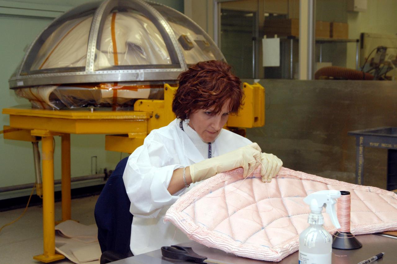 KENNEDY SPACE CENTER, FLA. -- In the Thermal Protection System Facility, Pilar Ryan, with United Space Alliance, stitches a piece of insulation blanket for Atlantis. The blankets will be sewn into the inside of a ring that will be inserted in the nose cap. In the background is a cover for the nose cap. The blankets consist of layered, pure silica felt sandwiched between a layer of silica fabric (the hot side) and a layer of S-Glass fabric. The blankets are semi-rigid and can be made as large as 30 inches by 30 inches. The blanket is through-stitched with pure silica thread in a 1-inch grid pattern. After fabrication, the blanket is bonded directly to the vehicle structure and finally coated with a high purity silica coating that improves erosion resistance.