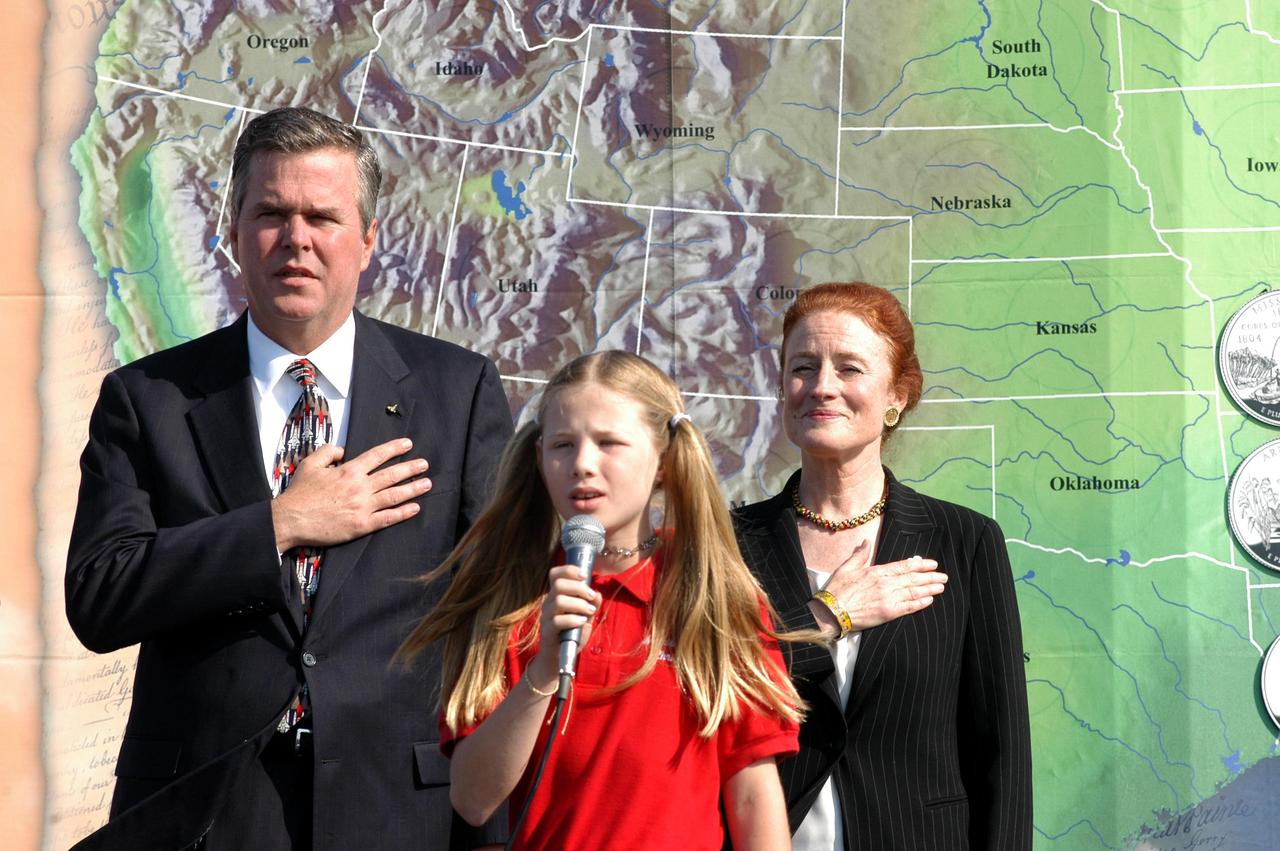 KENNEDY SPACE CENTER, FLA. -- During opening ceremonies at the KSC Visitor Complex launching the new Florida quarter, Florida Gov. Jeb Bush (left) and U.S. Mint Director Henrietta Holsman Fore (right) stand at attention while fourth grader Alexandra Schenck, from Merritt Island Christian School, sings the national anthem. Also participating in the event were NASA Administrator Sean O’Keefe and Deputy Secretary of the Treasury Samuel W. Bodman.  Center Director Jim Kennedy emceed the ceremonies. .  The quarter celebrates Florida as a destination for explorers in the past, a launch site for future explorers into space and an inviting place for visitors today.