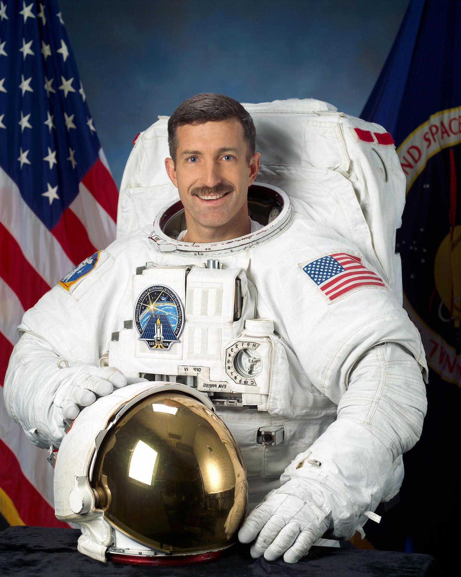 A man wearing a spacesuit smiles and stands in front of the American flag. He is holding his helmet in front of himself.