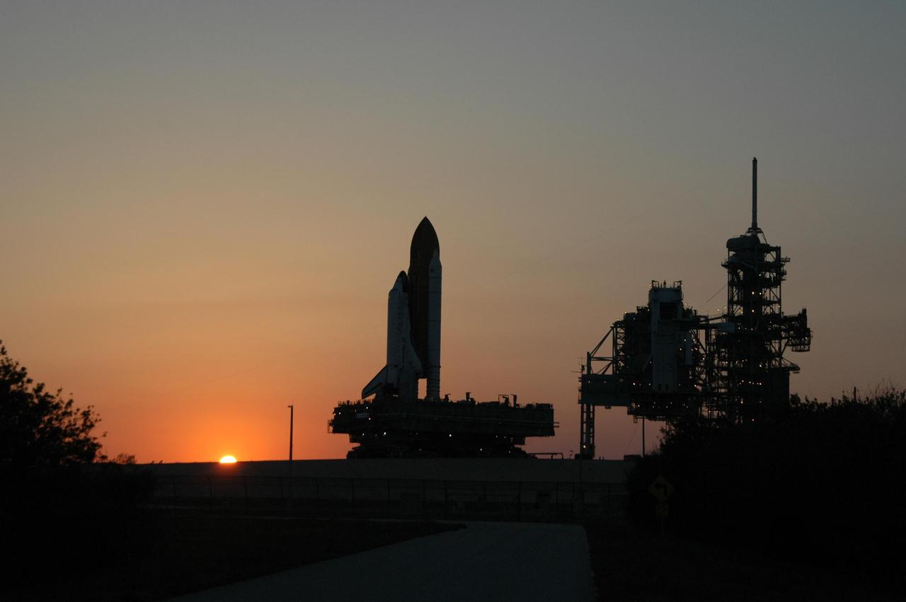 KENNEDY SPACE CENTER, FLA. -- The sun is setting as Space Shuttle Discovery arrives on the hardstand of Launch Pad 39B at NASA's Kennedy Space Center. At right are the fixed and rotating service structures. First motion was at 12:45 p.m. EDT. The shuttle rests on a mobile launcher platform and made the 4.2-mile journey from the Vehicle Assembly Building via the crawler-transporter beneath the platform. The rollout is an important step before launch of Discovery on mission STS-121 to the International Space Station. First motion of the shuttle leaving NASA's Vehicle Assembly Building was at 12:45 p.m. EDT. Discovery's launch is targeted for July 1 in a launch window that extends to July 19. During the 12-day mission, Discovery's crew will test new hardware and techniques to improve shuttle safety, as well as deliver supplies and make repairs to the station. Photo credit: NASA/Ken Thornsley