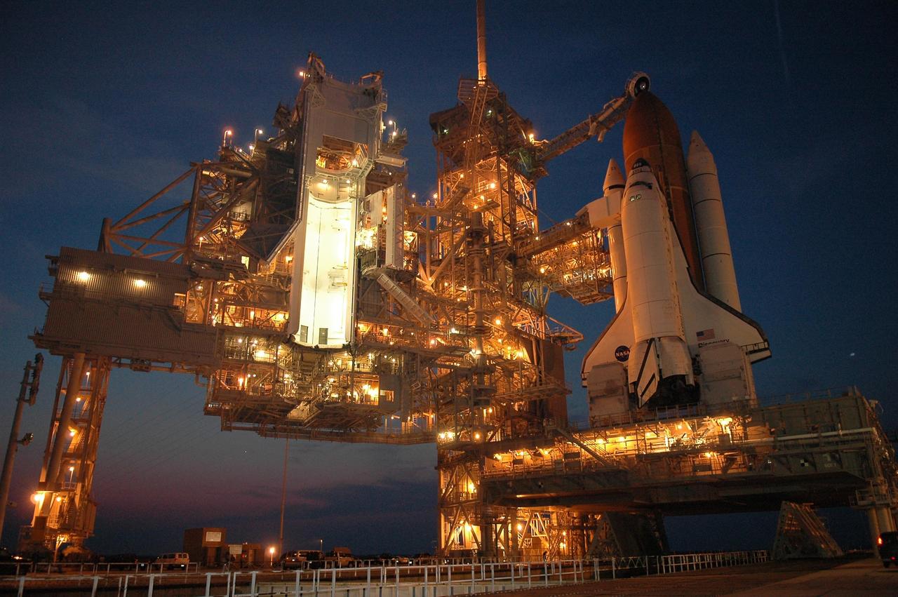 KENNEDY SPACE CENTER, FLA. - Against a dimming sky after sunset, lights on the structures on Launch Pad 39B cast a warm glow on Space Shuttle Discovery. The rotating service structure at left has been rolled back in preparation for launch July 1 on mission STS-121. Extending toward the cockpit of the shuttle is the orbiter access arm with the White Room extended. The White Room provides access into the orbiter for the astronauts. Above the golden external tank is the vent hood (known as the "beanie cap") at the end of the gaseous oxygen vent arm. Vapors are created as the liquid oxygen in the external tank boil off. The hood vents the gaseous oxygen vapors away from the space shuttle vehicle. The RSS provides protected access to the orbiter for changeout and servicing of payloads at the pad. The structure is supported by a rotating bridge that pivots about a vertical axis on the west side of the pad's flame trench. The hinge column rests on the pad surface and is braced to the fixed service structure. Support for the outer end of the bridge is provided by two eight-wheel, motor-driven trucks that move along circular twin rails installed flush with the pad surface. The track crosses the flame trench on a permanent bridge. The RSS is 102 feet long, 50 feet wide and 130 feet high. The structure has orbiter access platforms at five levels to provide access to the payload bay while the orbiter is being serviced in the RSS. Each platform has independent extendable planks that can be arranged to conform to a payload's configuration. This mission is the 115th shuttle flight and the 18th U.S. flight to the International Space Station. Photo credit: NASA/Kim Shiflett