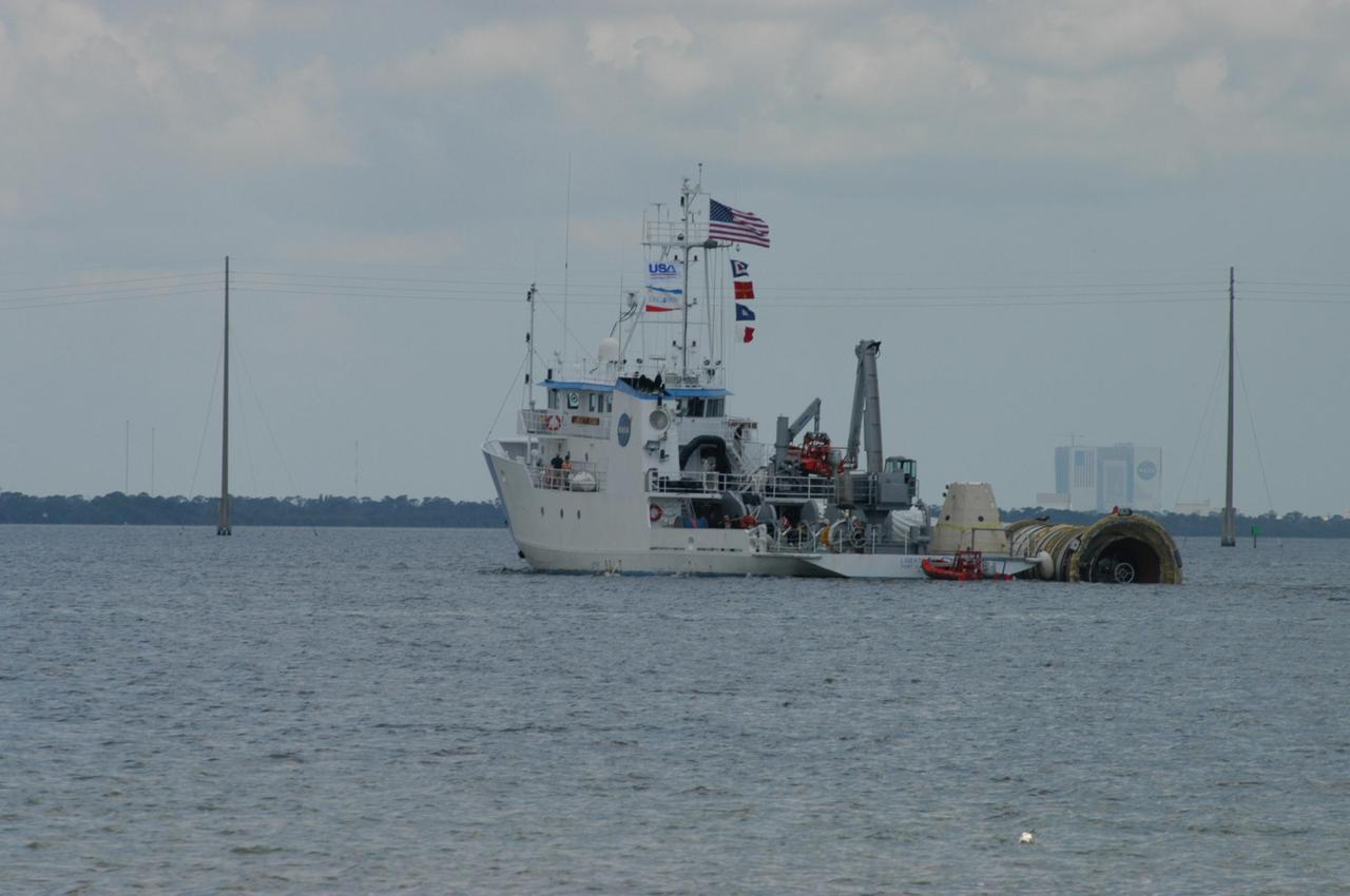KENNEDY SPACE CENTER, FLA. - With the Vehicle Assembly Building in the background, the SRB Retrieval Ship Liberty Star nears Cape Canaveral Air Force Station with a spent solid rocket booster alongside. The booster is from Space Shuttle Discovery, which launched on July 4. The space shuttle’s solid rocket booster casings and associated flight hardware are recovered at sea. The boosters impact the Atlantic Ocean approximately seven minutes after liftoff. The splashdown area is a square of about 6 by 9 nautical miles located about 140 nautical miles downrange from the launch pad. The retrieval ships are stationed approximately 8 to 10 nautical miles from the impact area at the time of splashdown. As soon as the boosters enter the water, the ships accelerate to a speed of 15 knots and quickly close on the boosters. The pilot chutes and main parachutes are the first items to be brought on board. With the chutes and frustum recovered, attention turns to the boosters. The ship’s tow line is connected and the booster is returned to the Port and ,after transfer to a position alongside the ship, to Hangar AF at Cape Canaveral Air Force Station. There, the expended boosters are disassembled, refurbished and reloaded with solid propellant for reuse. Photo credit: NASA/George Shelton