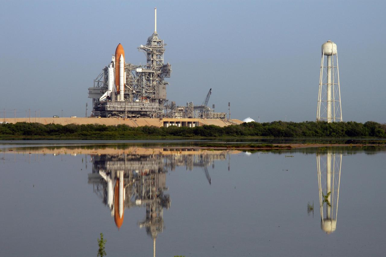 KENNEDY SPACE CENTER, FLA. -    Reflected in the nearby pool of water, Space Shuttle Atlantis arrives on the hard stand on Launch Pad 39B, propelled by the crawler-transporter.  At right is the 290-foot high, 300,000-gallon water tank that aids in sound suppression during launch. The water releases just prior to the ignition of the shuttle engines and flows through 7-foot-diameter pipes for about 20 seconds, pouring into 16 nozzles atop the flame deflectors and from outlets in the main engines exhaust hole in the mobile launcher platform. Atop the fixed service structure is the 80-foot lightning mast that helps provide lightning protection. The slow speed of the crawler results in a 6- to 8-hour trek to the pad approximately 4 miles away.  Atlantis' launch window begins Aug. 27 for an 11-day mission to the International Space Station. The STS-115 crew of six astronauts will continue construction of the station and install their cargo, the Port 3/4 truss segment with its two large solar arrays.   Photo credit: NASA/Tony Gray