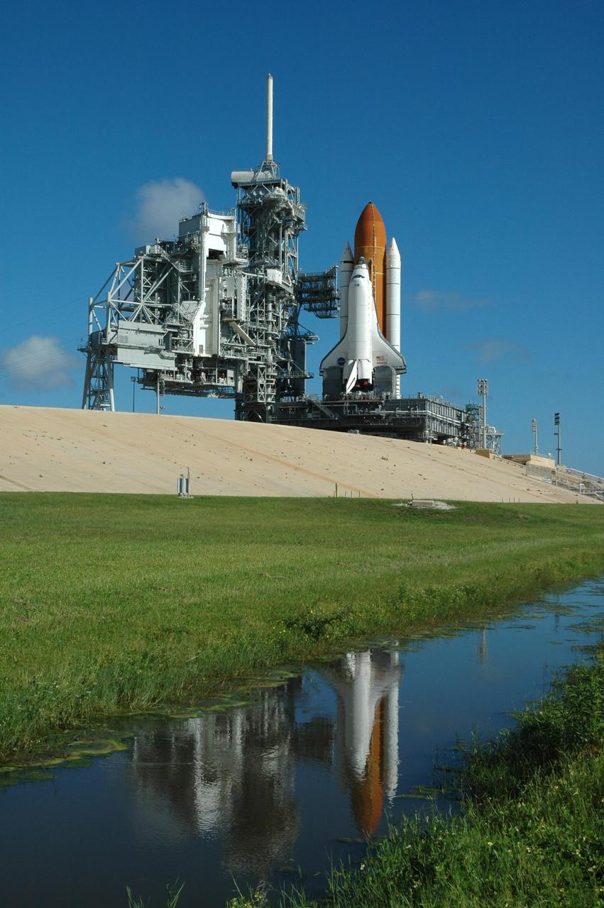 KENNEDY SPACE CENTER, FLA. - Space Shuttle Atlantis is reflected in the water near Launch Pad 39B. The rotating service structure has been rolled away in anticipation of the rollback of the shuttle and mobile launcher platform to the Vehicle Assembly Building (VAB), approximately 4 miles away. Despite the clear blue skies, the rollback is a safety precaution as the area waits for the arrival of Tropical Storm Ernesto. The storm is forecast to be bringing 58-mph to 70-mph winds in the next 24 hours. The shuttle will be moved into high bay 2, on the southwest side of the VAB, for protection from the storm. Photo credit: NASA/Jim Grossmann