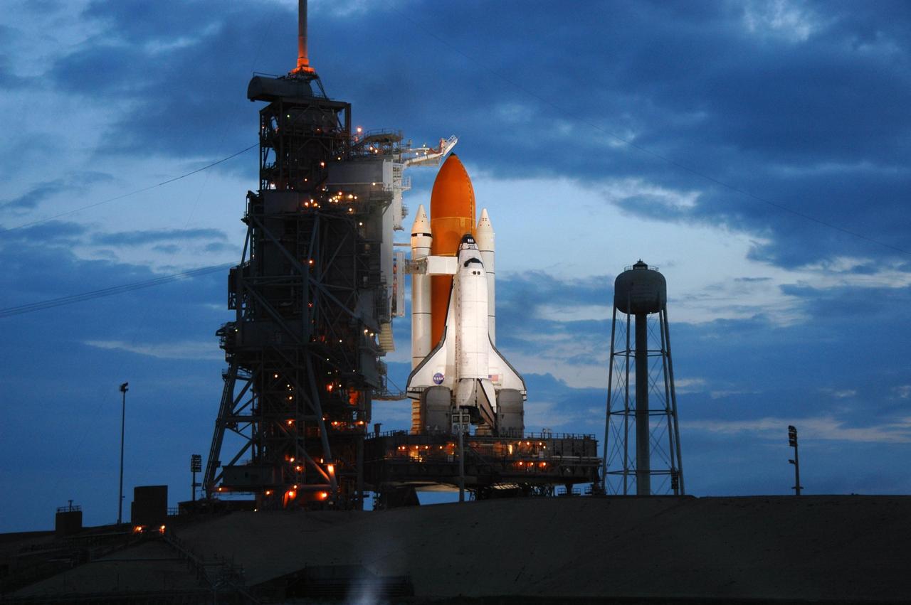 KENNEDY SPACE CENTER, FLA. -   The morning sky lightens behind Space Shuttle Atlantis while lights on the fixed service structure (FSS)  still illuminate the orbiter on Launch Pad 39B. Atlantis was originally scheduled to launch at 12:29 p.m. EDT on this date, but  a 24-hour scrub was called by mission managers due to a concern with Fuel Cell 1.  Seen poised above the orange external tank is the vent hood (known as the "beanie cap") at the end of the gaseous oxygen vent arm. Vapors are created as the liquid oxygen in the external tank boil off. The hood vents the gaseous oxygen vapors away from the space shuttle vehicle. Extending from the FSS to Atlantis is the orbiter access arm with the White Room at the end.  The White Room provides entry into the orbiter through the hatch.  At right is the 300,000-gallon water tank that releases its contents onto the mobile launcher platform during liftoff to aid sound suppression. During the STS-115 mission, Atlantis' astronauts will deliver and install the 17.5-ton, bus-sized P3/P4 integrated truss segment on the station. The girder-like truss includes a set of giant solar arrays, batteries and associated electronics and will provide one-fourth of the total power-generation capability for the completed station. This mission is the 116th space shuttle flight, the 27th flight for orbiter Atlantis, and the 19th U.S. flight to the International Space Station. STS-115 is scheduled to last 11 days with a planned landing at KSC.   Photo credit: NASA/Troy Cryder