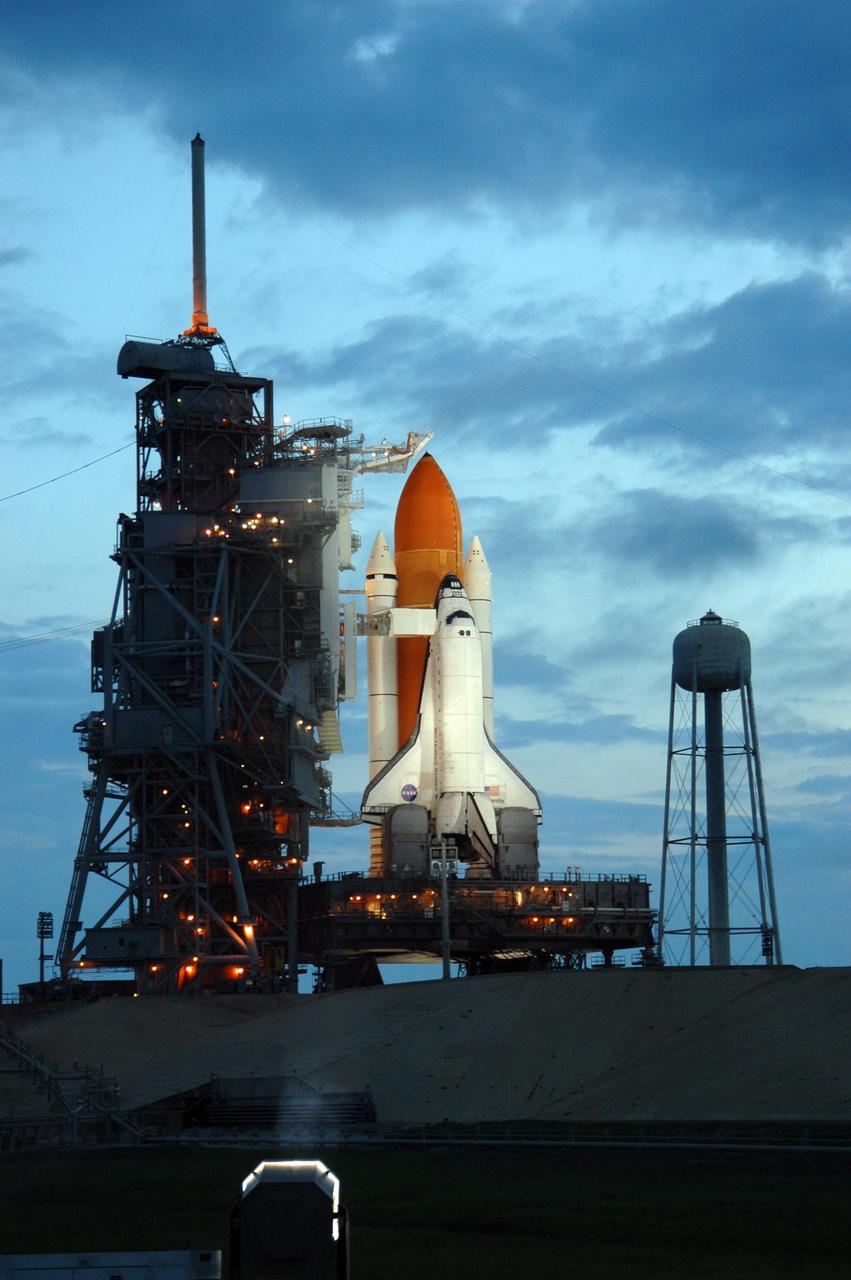 KENNEDY SPACE CENTER, FLA. -   The morning sky lightens behind Space Shuttle Atlantis while lights on the fixed service structure (FSS)  still illuminate the orbiter on Launch Pad 39B. Atlantis was originally scheduled to launch at 12:29 p.m. EDT on this date, but  a 24-hour scrub was called by mission managers due to a concern with Fuel Cell 1.  Seen poised above the orange external tank is the vent hood (known as the "beanie cap") at the end of the gaseous oxygen vent arm. Vapors are created as the liquid oxygen in the external tank boil off. The hood vents the gaseous oxygen vapors away from the space shuttle vehicle. Extending from the FSS to Atlantis is the orbiter access arm with the White Room at the end.  The White Room provides entry into the orbiter through the hatch.  At right is the 300,000-gallon water tank that releases its contents onto the mobile launcher platform during liftoff to aid sound suppression.  During the STS-115 mission, Atlantis' astronauts will deliver and install the 17.5-ton, bus-sized P3/P4 integrated truss segment on the station. The girder-like truss includes a set of giant solar arrays, batteries and associated electronics and will provide one-fourth of the total power-generation capability for the completed station. This mission is the 116th space shuttle flight, the 27th flight for orbiter Atlantis, and the 19th U.S. flight to the International Space Station. STS-115 is scheduled to last 11 days with a planned landing at KSC.   Photo credit: NASA/Troy Cryder