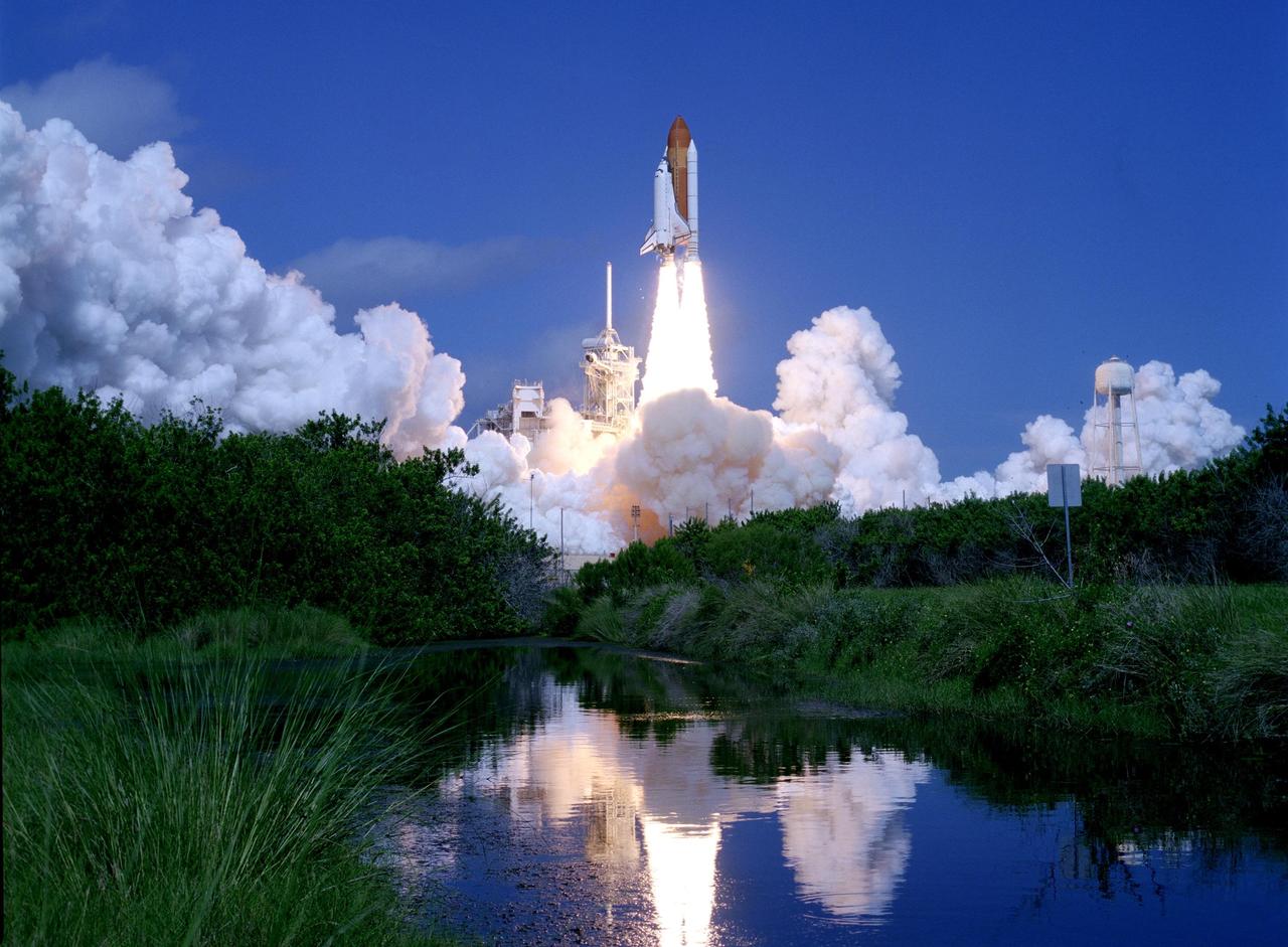 KENNEDY SPACE CENTER, FLA. -    Launch of Space Shuttle Atlantis into the deep blue sky is reflected in the nearby water framed by the green underbrush.  Clouds of smoke and steam spread side to side across Launch Pad 39B.  Atlantis is heading for a rendezvous with the International Space Station on mission STS-115.  Liftoff was on-time at 11:14:55 a.m. EDT. After several launch attempts were scrubbed due to weather and technical concerns, this launch was executed perfectly. Mission STS-115 is the 116th space shuttle flight, the 27th flight for orbiter Atlantis, and the 19th U.S. flight to the International Space Station.  During the mission, Atlantis' astronauts will deliver and install the 17.5-ton, bus-sized P3/P4 integrated truss segment on the station. The girder-like truss includes a set of giant solar arrays, batteries and associated electronics and will provide one-fourth of the total power-generation capability for the completed station. STS-115 is scheduled to last 11 days with a planned landing at KSC