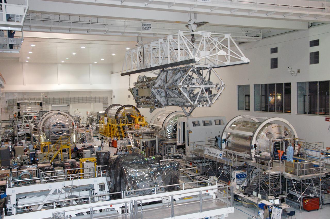 In the Space Station Processing Facility, an overhead crane moves the S3/S4 integrated truss above the floor to a payload canister. Several space station modules can be seen at various points on the floor. After it is stowed in the canister, the S3/S4 truss will be transported to the launch pad. The truss is the payload on mission STS-117, targeted for launch on March 15.