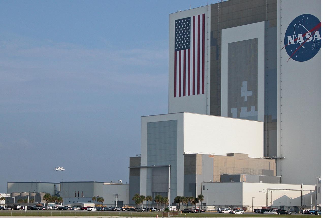 Through broken clouds, the shuttle carrier aircraft, or SCA, and its passenger Atlantis ease their way past the Vehicle Assembly Building, at right, for a landing at the KSC Shuttle Landing Facility, known as the SLF. The aircraft is a modified Boeing 747 jetliner. Atlantis landed at Edwards Air Force Base in California to end mission STS-117. The return to KSC began July 1 and took three days after stops across the country for fuel. The last stop was at Ft. Campbell in Kentucky. Weather conditions over the last leg postponed the return trip until July 3. Touchdown was at 8:27 a.m. EDT. Atlantis will be removed from the back of the SCA via the mate/demate device at the SLF. It will then be towed to the Orbiter Processing Facility to begin processing for its next launch, mission STS-122 in December.