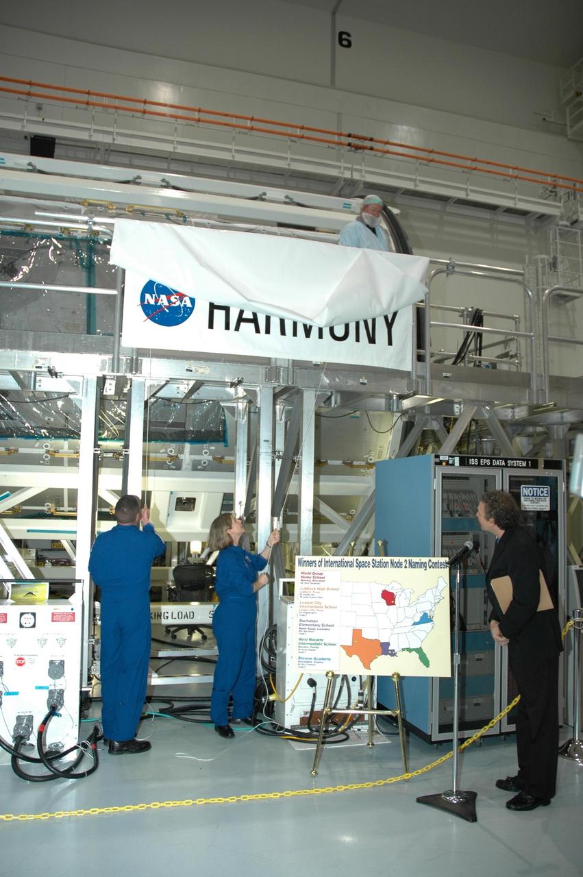 KENNEDY SPACE CENTER, FLA. -- In the Space Station Processing Facility, Mission STS-120 Pilot George Zamka (left) and Commander Pam Melroy begin to unveil the Node 2 module's new name, Harmony, as Russ Romanella, director of International Space Station and Spacecraft Processing presides over the ceremony. The name, Harmony, was chosen from an academic competition involving thousands of students in kindergarten through high school. The Node 2 Challenge required students to learn about the International Space Station, build a scale model of the module, and write an essay explaining their proposed name. This will be the first U.S. piece of the space station named by someone other than a NASA official. Node 2 is a pressurized module that will act as a connecting port and passageway to additional international science labs and supply spacecraft. It also will be a work platform for the station's robotic arm. The module is scheduled to fly on mission STS-120 aboard Space Shuttle Atlantis targeted for later this year. Photo credit: NASA/Jim Grossmann