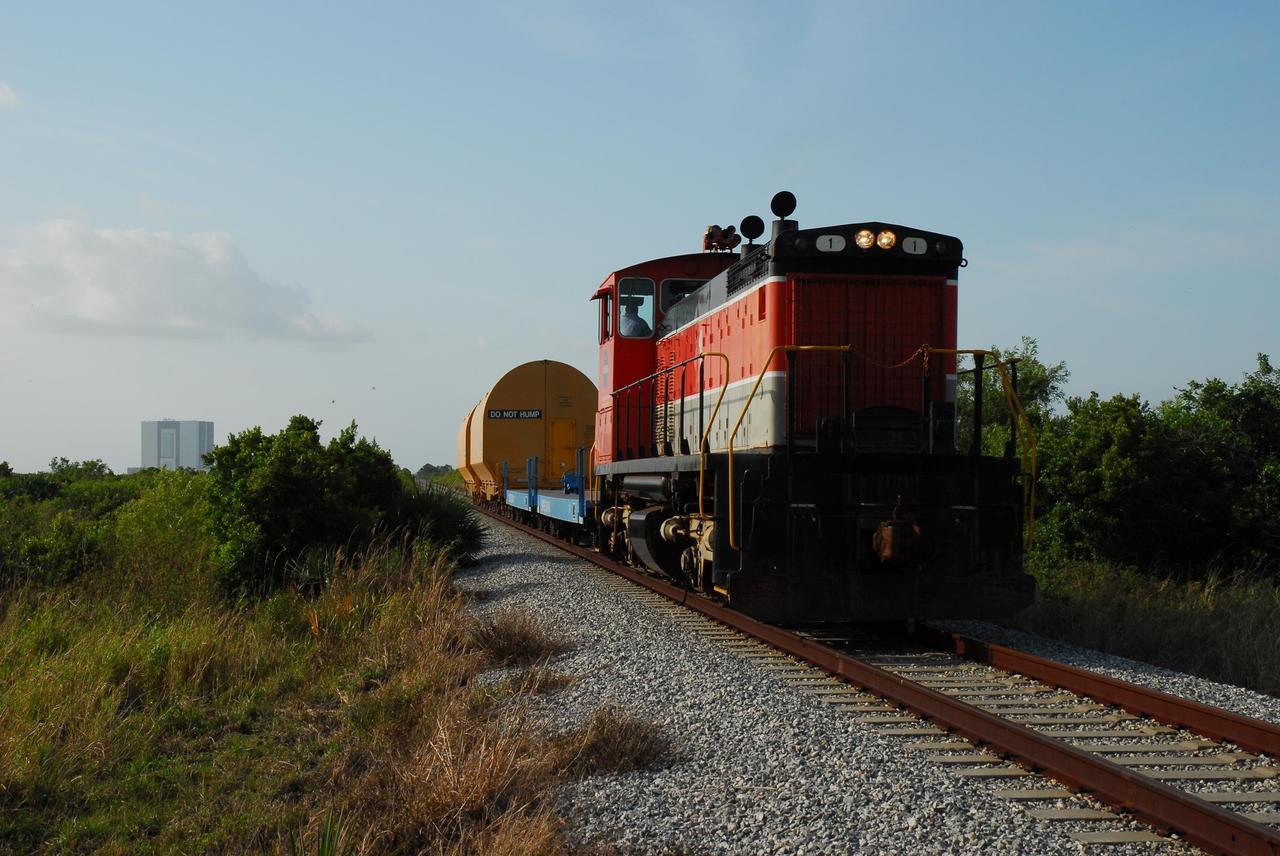 KENNEDY SPACE CENTER, FLA. --   The locomotive and rail cars carrying solid rocket booster motor segments and two aft exit cone segments roll to the Rotation, Processing and Surge Facility (RPSF) in Kennedy Space Center's Launch Complex 39 Area.  In the background, at left, is the Vehicle Assembly Building.  The RPSF is used for solid rocket motor receiving, rotation and inspection, and supports aft booster buildup.  When live solid rocket motor segments arrive at the processing facility, they  are positioned under one of the cranes. Handling slings are then attached to and remove the railcar cover. The segment is inspected while it remains horizontal.  The two overhead cranes hoist the segment, rotate it to a vertical position and place it on a fixed stand. The aft handling ring is then removed. The segment is hoisted again and lowered onto a transportation and storage pallet, and the forward handling ring is removed to allow inspections. It is then transported to one of the surge buildings and temporarily stored until it is needed for booster stacking in the VAB.  While enroute, solid rocket motor segments were involved in a derailment in Alabama.  The rail cars carrying these segments remained upright and were undamaged.  An inspection determined these segment cars could continue on to Florida.  The segments themselves will undergo further evaluation at Kennedy before they are cleared for flight.  Other segments involved in the derailment will be returned to a plant in Utah for further evaluation.  Photo credit: NASA/George Shelton