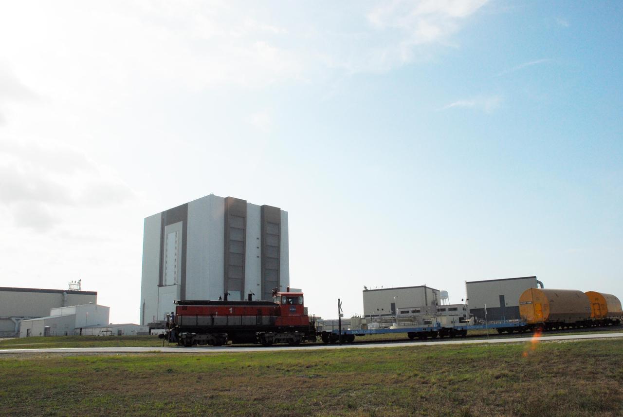 KENNEDY SPACE CENTER, FLA. --   The locomotive and rail cars carrying solid rocket booster motor segments and two aft exit cone segments roll past the Vehicle Assembly Building to the Rotation, Processing and Surge Facility (RPSF) in Kennedy Space Center's Launch Complex 39 Area.  The RPSF is used for solid rocket motor receiving, rotation and inspection, and supports aft booster buildup.  When live solid rocket motor segments arrive at the processing facility, they  are positioned under one of the cranes. Handling slings are then attached to and remove the railcar cover. The segment is inspected while it remains horizontal.  The two overhead cranes hoist the segment, rotate it to a vertical position and place it on a fixed stand. The aft handling ring is then removed. The segment is hoisted again and lowered onto a transportation and storage pallet, and the forward handling ring is removed to allow inspections. It is then transported to one of the surge buildings and temporarily stored until it is needed for booster stacking in the VAB.  While enroute, solid rocket motor segments were involved in a derailment in Alabama.  The rail cars carrying these segments remained upright and were undamaged.  An inspection determined these segment cars could continue on to Florida.  The segments themselves will undergo further evaluation at Kennedy before they are cleared for flight.  Other segments involved in the derailment will be returned to a plant in Utah for further evaluation.  Photo credit: NASA/George Shelton