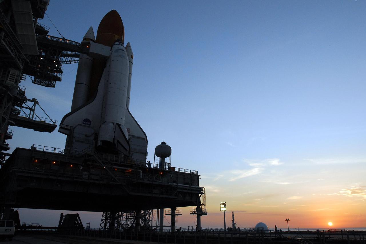 KENNEDY SPACE CENTER, FLA. -- The rising sun, at right, reveals Space Shuttle Endeavour sitting on Launch Pad 39A. First motion out of the Vehicle Assembly Building was at 8:10 p.m. July 10, and the shuttle was hard down on the pad at 3:02 a.m. July 11. On the left is the fixed service structure with the orbiter access arm already extended to the orbiter. The top of the 290-foot-tall water tank is seen to the right of the shuttle. It provides the deluge over the mobile launcher platform for sound suppression during liftoff. Endeavour is scheduled to launch on mission STS-118 on Aug. 7. During the mission, Endeavour will carry into orbit the S5 truss, SPACEHAB module and external stowage platform 3. The mission is the 22nd flight to the International Space Station and will mark the first flight of Mission Specialist Barbara Morgan, the teacher-turned-astronaut whose association with NASA began more than 20 years ago. STS-118 will be the first flight since 2002 for Endeavour, which has undergone extensive modifications, including the addition of safety upgrades already added to orbiters Discovery and Atlantis. Photo credit: NASA/George Shelton