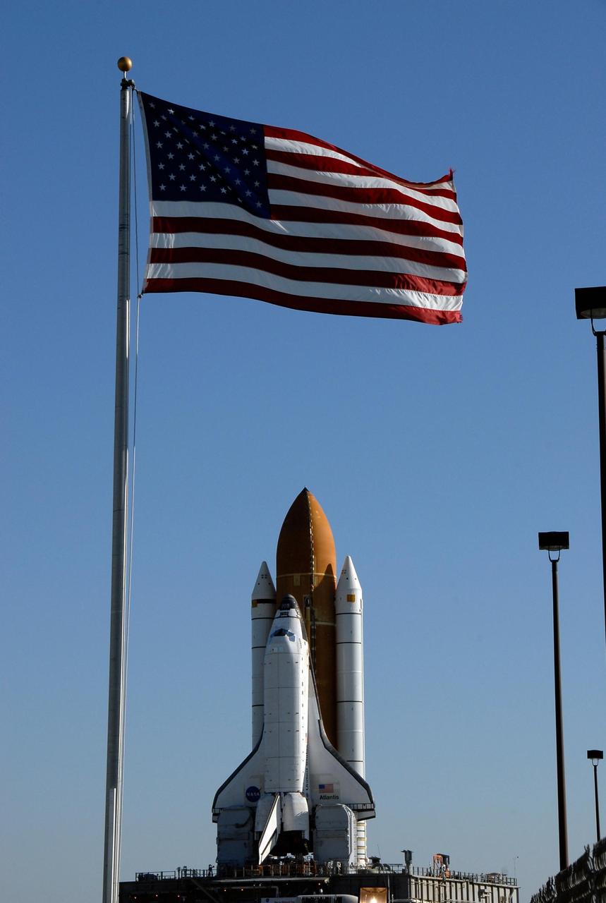 KENNEDY SPACE CENTER, FLA. -- Two symbols of American pride, the Space Shuttle Atlantis and a United States flag, appear side by side as Atlantis makes its way to Launch Pad 39A on a breezy Florida morning. The crawler transporter has a top speed of one mile per hour while it is moving the space shuttle. First motion out of the Vehicle Assembly Building was at 4:43 a.m. EST.  Rollout is a milestone for Atlantis' launch to the International Space Station on mission STS-122, targeted for Dec. 6. On this mission, Atlantis will deliver the Columbus module to the International Space Station. The European Space Agency's largest contribution to the station, Columbus is a multifunctional, pressurized laboratory that will be permanently attached to U.S. Node 2, called Harmony. The module is approximately 23 feet long and 15 feet wide, allowing it to hold 10 large racks of experiments. The laboratory will expand the research facilities aboard the station, providing crew members and scientists from around the world the ability to conduct a variety of experiments in the physical, materials and life sciences.   Photo credit: NASA/Kim Shiflett
