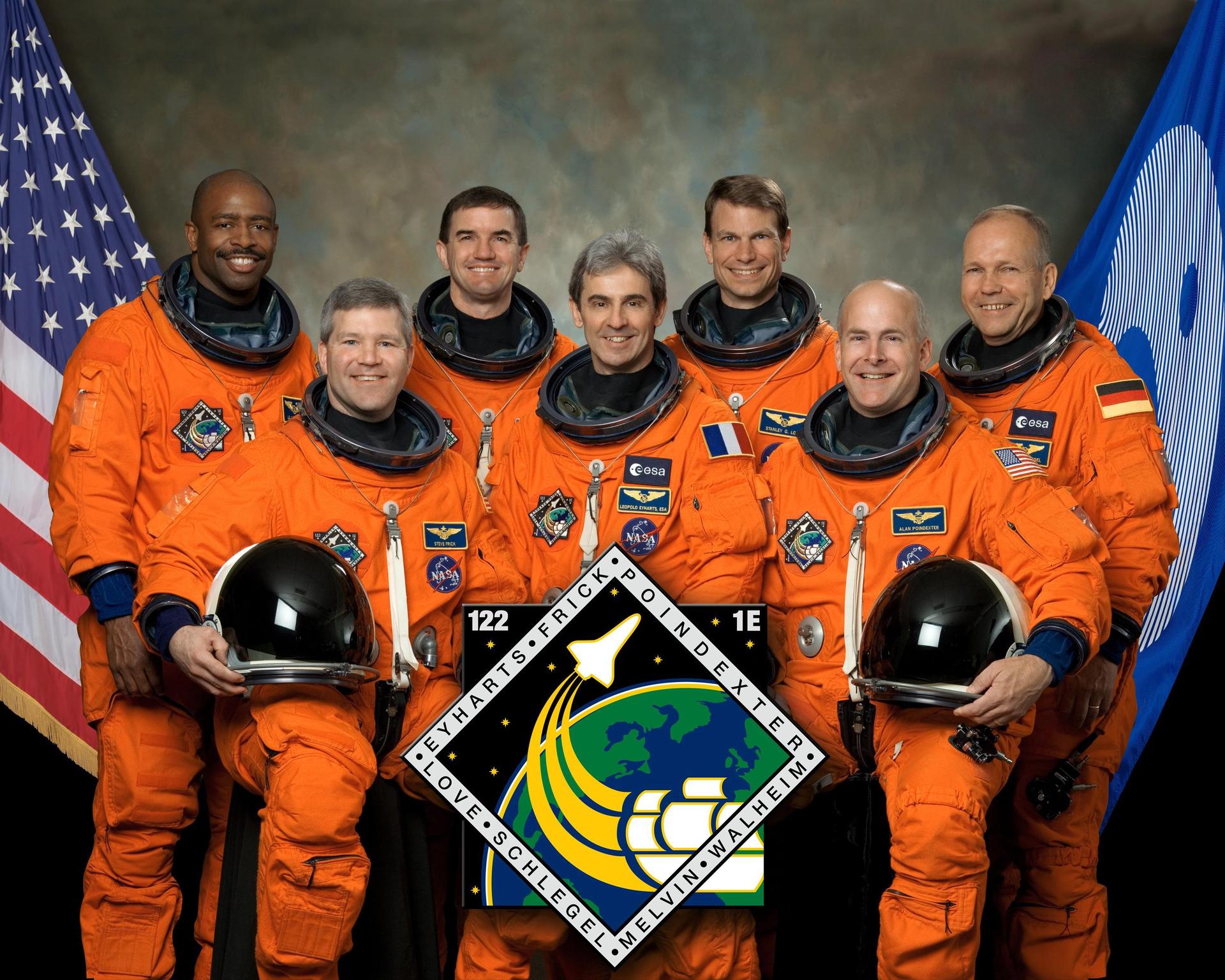 Crew members of STS-122 in orange flight suits pose for crew photo in front of flags.