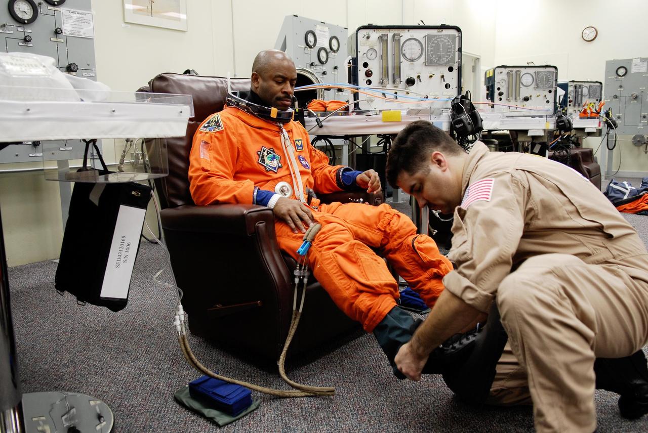 KENNEDY SPACE CENTER, FLA. -- A suit technician helps space shuttle Atlantis STS-122 Mission Specialist Leland Melvin put on his boots, part of his launch and entry suit, in astronaut crew quarters. The STS-122 crew is preparing for a simulated launch countdown aboard Atlantis, part of terminal countdown demonstration test, or TCDT, activities at NASA's Kennedy Space Center. The TCDT is a dress rehearsal for launch and also provides astronauts and ground crews with equipment familiarization and emergency egress training. On mission STS-122, Atlantis will deliver the Columbus module to the International Space Station. The European Space Agency's largest single contribution to the station, Columbus is a multifunctional, pressurized laboratory that will be permanently attached to U.S. Node 2, called Harmony. The laboratory will expand the research facilities aboard the station, providing crew members and scientists from around the world the ability to conduct a variety of experiments in the physical, materials and life sciences. Launch is targeted for Dec. 6. Photo credit: NASA/Kim Shiflett