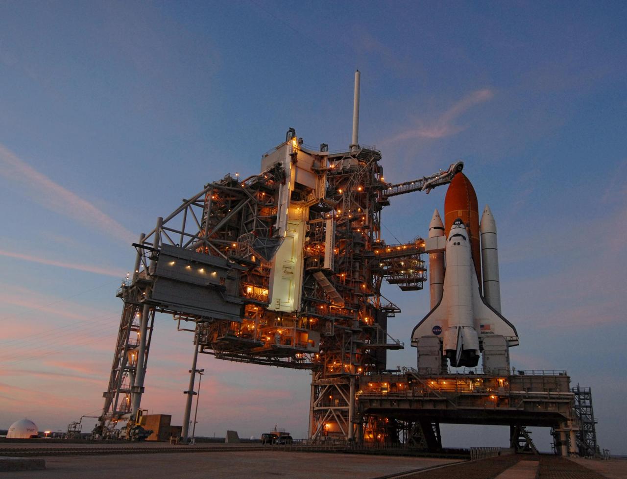 KENNEDY SPACE CENTER, FLA. --  Against a sunset sky streaked with pink, lights on the fixed and rotating service structures illuminate Launch Pad 39A where space shuttle Atlantis is still poised after its launch on mission STS-122 was postponed Thursday.  Shuttle program managers decided at 9:56 a.m. to postpone the launch because of an issue with a fuel cut-off sensor system inside the external fuel tank. This is one of several systems that protect the shuttle's main engines by triggering their shut down if fuel runs unexpectedly low. During countdown activities this morning, two sensors failed a routine prelaunch check. There are four engine cut-off, or ECO, sensors inside the liquid hydrogen section of the tank, and Launch Commit Criteria require three of the four sensor systems to be functioning properly. The tank's liquid oxygen and liquid hydrogen was drained from the tank, and preparations will begin for a possible launch attempt Friday. NASA's launch rules have a preplanned procedure that states in the case of ECO sensor system failure, engineers need to drain the tank and verify all the sensors are working as they go dry. Atlantis carries the Columbus Laboratory, the European Space Agency's largest contribution to the construction of the space station.  When permanently attached to Node 2, the laboratory will carry out experiments in materials science, fluid physics and biosciences, as well as perform a number of technological applications, in a microgravity environment.  Photo credit: NASA/George Shelton
