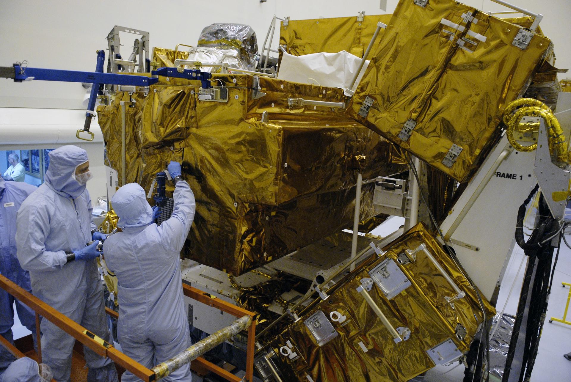 Image shows a gold protective layer covering NASA's Hubble Space Telescope at NASA Kennedy. People dressed in white and blue protective suits to not contaminate the telescope ahead of launch. Photo credit: NASA/Kim Shiflett
