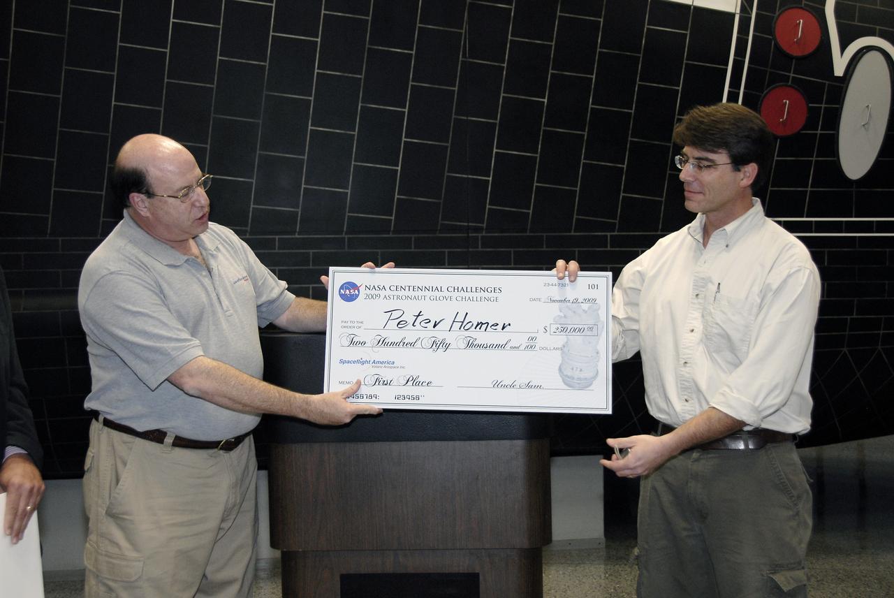 CAPE CANAVERAL, Fla. – Alan Hayes, left, chairman of Volanz Aerospace Inc., presents Peter Homer of Southwest Harbor, Maine, with the first place prize of $250,000 at the 2009 Astronaut Glove Challenge, part of NASA’s Centennial Challenges Program, at the Astronaut Hall of Fame near NASA’s Kennedy Space Center in Florida.    The nationwide competition focused on developing improved pressure suit gloves for astronauts to use while working in space.  During the challenge, the gloves were submitted to burst tests, joint force tests and tests to measure their dexterity and strength during operation in a glove box which simulates the vacuum of space.  Centennial Challenges is NASA’s program of technology prizes for the citizen-inventor. The winning prize for the Glove Challenge is $250,000 provided by the Centennial Challenges Program.  Photo credit: NASA/Kim Shiflett