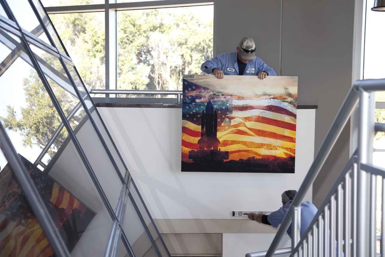CAPE CANAVERAL, Fla. -- Workers hang artwork in the second-floor lobby of the Propellants North Administrative and Maintenance Facility at NASA's Kennedy Space Center in Florida. The artwork was produced by Greg Lee, a graphics specialist with Abacus Technology Corp., and features a silhouette of a shuttle, one of the most recognizable American icons, rolling out to Launch Complex 39. The environmentally friendly facility is slated to be NASA's second Platinum-rated by the U.S. Green Building Council's (USGBC) Leadership in Environmental and Energy Design (LEED) certification system. It will be the space agency's first net-zero facility, which means it will produce enough energy onsite from renewable sources to offset what it requires to operate. The facility consists of a two-story administrative building to house managers, mechanics and technicians who fuel spacecraft at Kennedy, and a single-story shop to store cryogenic fuel transfer equipment. Photo credit: NASA/Frankie Martin
