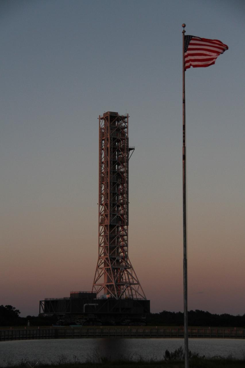 CAPE CANAVERAL, Fla. – The flag at NASA's Press Site is on guard as the mobile launcher, or ML, passes by the turn basin at NASA's Kennedy Space Center in Florida. The ML is nearing the end of its 4.2-mile trek from Launch Pad 39B to the park site near the Vehicle Assembly Building as the sun sets.    Data on the ML collected from structural and functional engineering tests during its two-week stay on the pad will be used in the next phases of construction. The 355-foot-tall ML structure, which took about two years to construct, will be modified by NASA’s 21st Century Ground Systems Program to support NASA’s Space Launch System, the heavy-lift rocket that will launch astronauts into deep space on future exploration missions. For more information, visit http://www.nasa.gov/exploration/systems/sls. Photo credit: NASA/Cory Huston