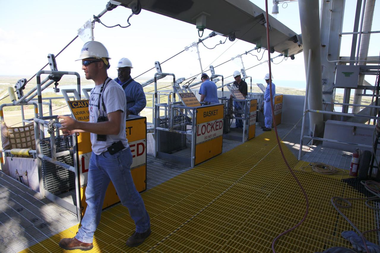 CAPE CANAVERAL, Fla. – At Launch Pad 39A at NASA’s Kennedy Space Center in Florida, technicians prepare for the final release of the seven slidewire baskets from the 195-foot level. After the baskets reach the ground they will be removed and put in storage.    The system of seven slidewire baskets at launch pads A and B provided an escape route for personnel inside the orbiter or on the orbiter access arm. The baskets are suspended from slidewires that extend from the pad’s Fixed Service Structure to a landing zone 1,200 feet to the west. Each basket could hold up to three people. A braking system catch net and drag chain slowed and then halted the baskets sliding down the wire approximately 55 miles per hour in about half a minute. Photo credit: NASA/Frankie Martin