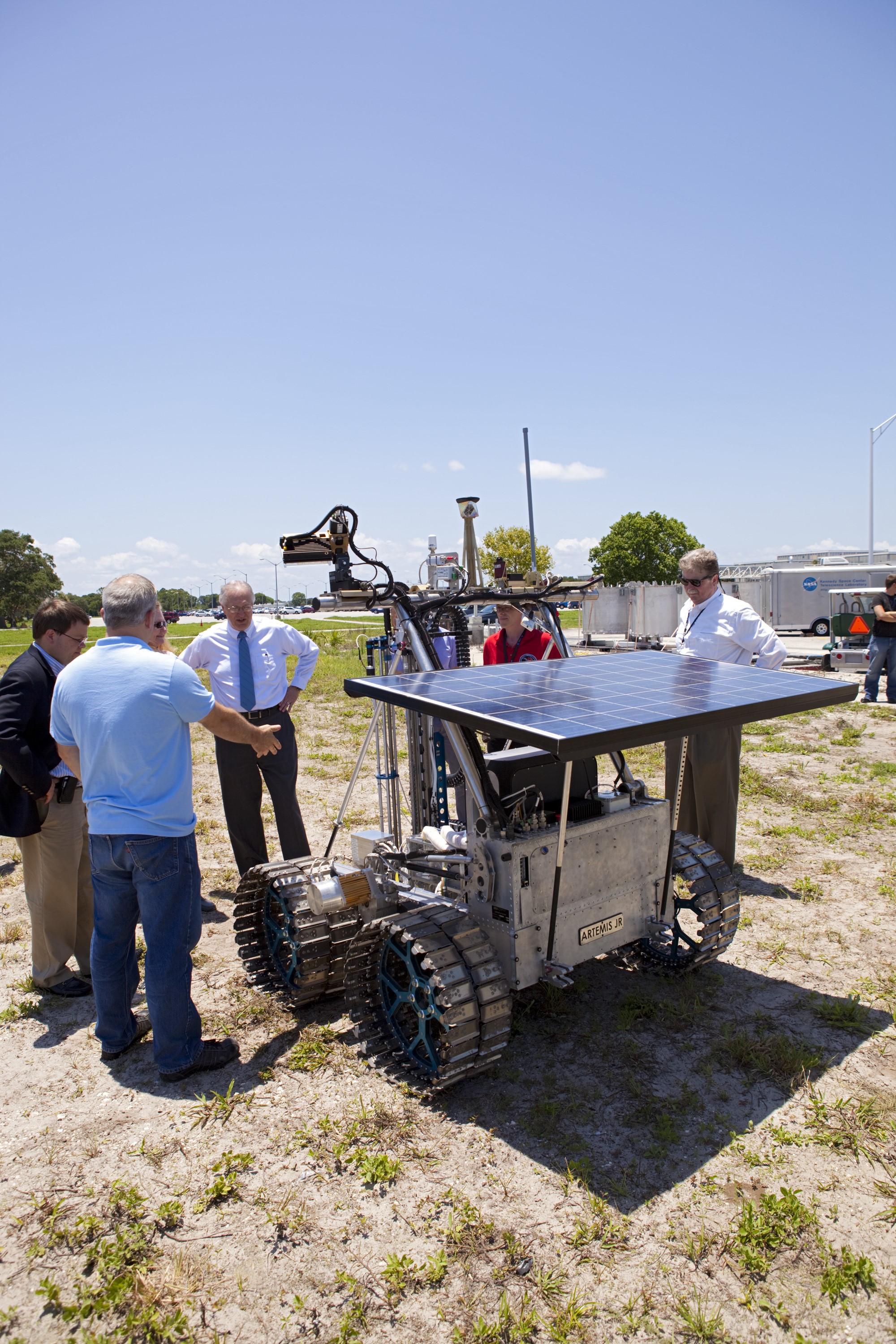 CAPE CANAVERAL, Fla. –  Media representatives discuss the design and operation of the prototype rover Artemis Jr. for NASA’s Regolith and Environment Science and Oxygen and Lunar Volatile Extraction, or RESOLVE, project with NASA In Situ Resource Utilization Project Manager William Larson, facing the rover, in a field beside the Operations and Checkout Building at NASA’s Kennedy Space Center in Florida.    The rover and its drill are provided by the Canadian Space Agency and work in concert with NASA science instruments to prospect for water, ice and other lunar resources.  RESOLVE also will demonstrate how future explorers can take advantage of resources at potential landing sites by manufacturing oxygen from soil. NASA will conduct field tests in July outside of Hilo, Hawaii, with equipment and concept vehicles that demonstrate how explorers might prospect for resources and make their own oxygen for survival while on other planetary bodies.  For more information, visit http://www.nasa.gov/exploration/analogs/index.html.  Photo credit: NASA/Dimitri Gerondidakis