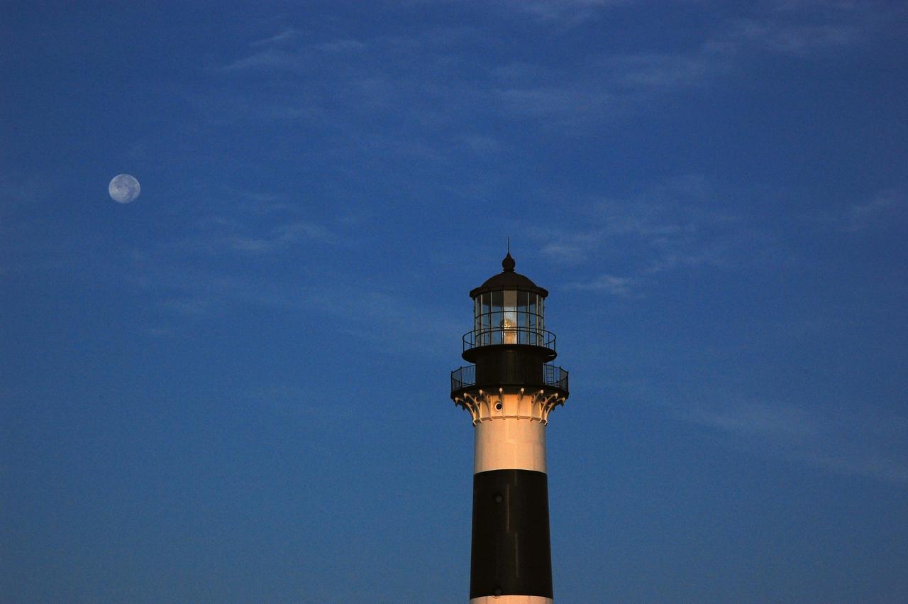 Cape Canaveral Air Force Station, Fla. -- The lantern room of the Cape Canaveral Lighthouse, with its modern first-order optic, takes on a warm glow as dawn breaks and a full moon still shines overhead. The Canaveral light is the only operating lighthouse owned by the U.S. Air Force. In 2000, the Coast Guard transferred ownership of the lighthouse structure and its grounds to the Air Force, which is now responsible for maintaining it. The U.S. Coast Guard continues to operate the beacon as an active navigational aid. The first lighthouse at Cape Canaveral was built near the tip of the Cape in 1848. The structure was only about 60 feet high with a rather dim light powered by whale oil. In 1859, work began nearby on a new, taller iron structure. Construction was halted during the Civil War, and the lighthouse finally was finished in 1868. The structure, with a brick lining inside its iron exterior, was painted with its "daymark" black and white horizontal bands in 1873 to make it easier to identify during the day as a navigation point. Between 1892 and 1894, the lighthouse was dismantled and moved to its new home about a mile from the coast, where it stands today. Photo credit: NASA/Ben Smegelsky