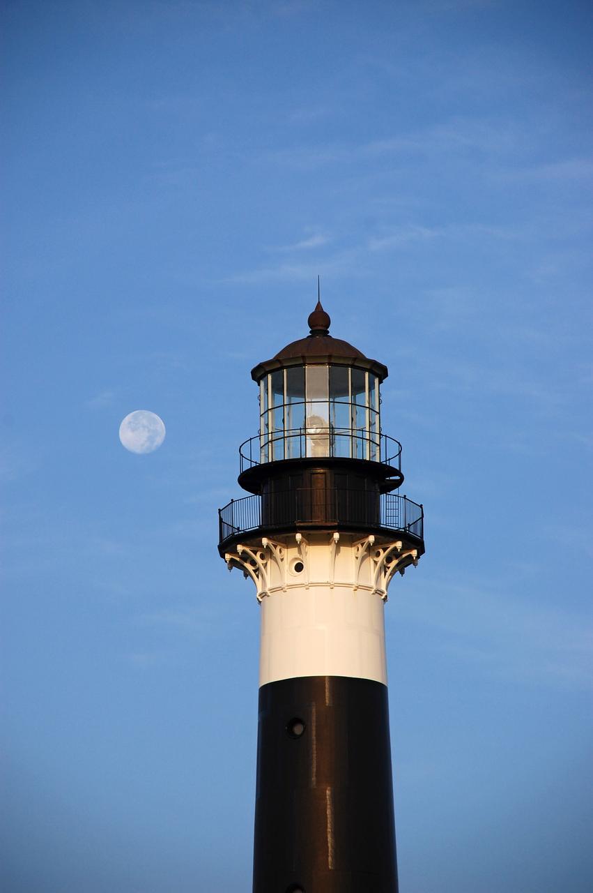 Cape Canaveral Air Force Station, Fla. -- The lantern room of the Cape Canaveral Lighthouse, with its modern first-order optic, takes on a warm glow as dawn breaks and a full moon still shines overhead. The Canaveral light is the only operating lighthouse owned by the U.S. Air Force. In 2000, the Coast Guard transferred ownership of the lighthouse structure and its grounds to the Air Force, which is now responsible for maintaining it. The U.S. Coast Guard continues to operate the beacon as an active navigational aid. The first lighthouse at Cape Canaveral was built near the tip of the Cape in 1848. The structure was only about 60 feet high with a rather dim light powered by whale oil. In 1859, work began nearby on a new, taller iron structure. Construction was halted during the Civil War, and the lighthouse finally was finished in 1868. The structure, with a brick lining inside its iron exterior, was painted with its "daymark" black and white horizontal bands in 1873 to make it easier to identify during the day as a navigation point. Between 1892 and 1894, the lighthouse was dismantled and moved to its new home about a mile from the coast, where it stands today. Photo credit: NASA/Ben Smegelsky