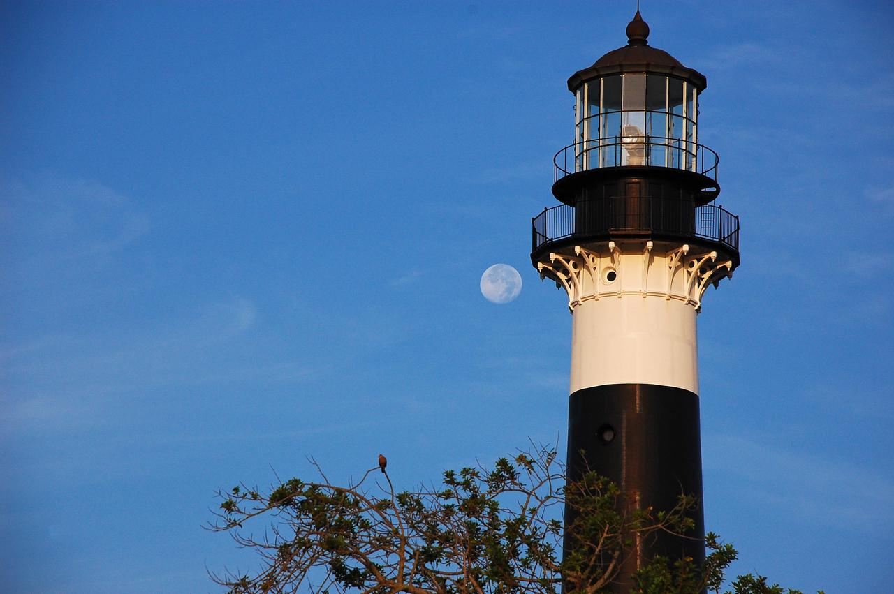 Cape Canaveral Air Force Station, Fla. -- The lantern room of the Cape Canaveral Lighthouse, with its modern first-order optic, takes on a warm glow as dawn breaks and a full moon still shines overhead. The Canaveral light is the only operating lighthouse owned by the U.S. Air Force. In 2000, the Coast Guard transferred ownership of the lighthouse structure and its grounds to the Air Force, which is now responsible for maintaining it. The U.S. Coast Guard continues to operate the beacon as an active navigational aid. The first lighthouse at Cape Canaveral was built near the tip of the Cape in 1848. The structure was only about 60 feet high with a rather dim light powered by whale oil. In 1859, work began nearby on a new, taller iron structure. Construction was halted during the Civil War, and the lighthouse finally was finished in 1868. The structure, with a brick lining inside its iron exterior, was painted with its "daymark" black and white horizontal bands in 1873 to make it easier to identify during the day as a navigation point. Between 1892 and 1894, the lighthouse was dismantled and moved to its new home about a mile from the coast, where it stands today. Photo credit: NASA/Ben Smegelsky