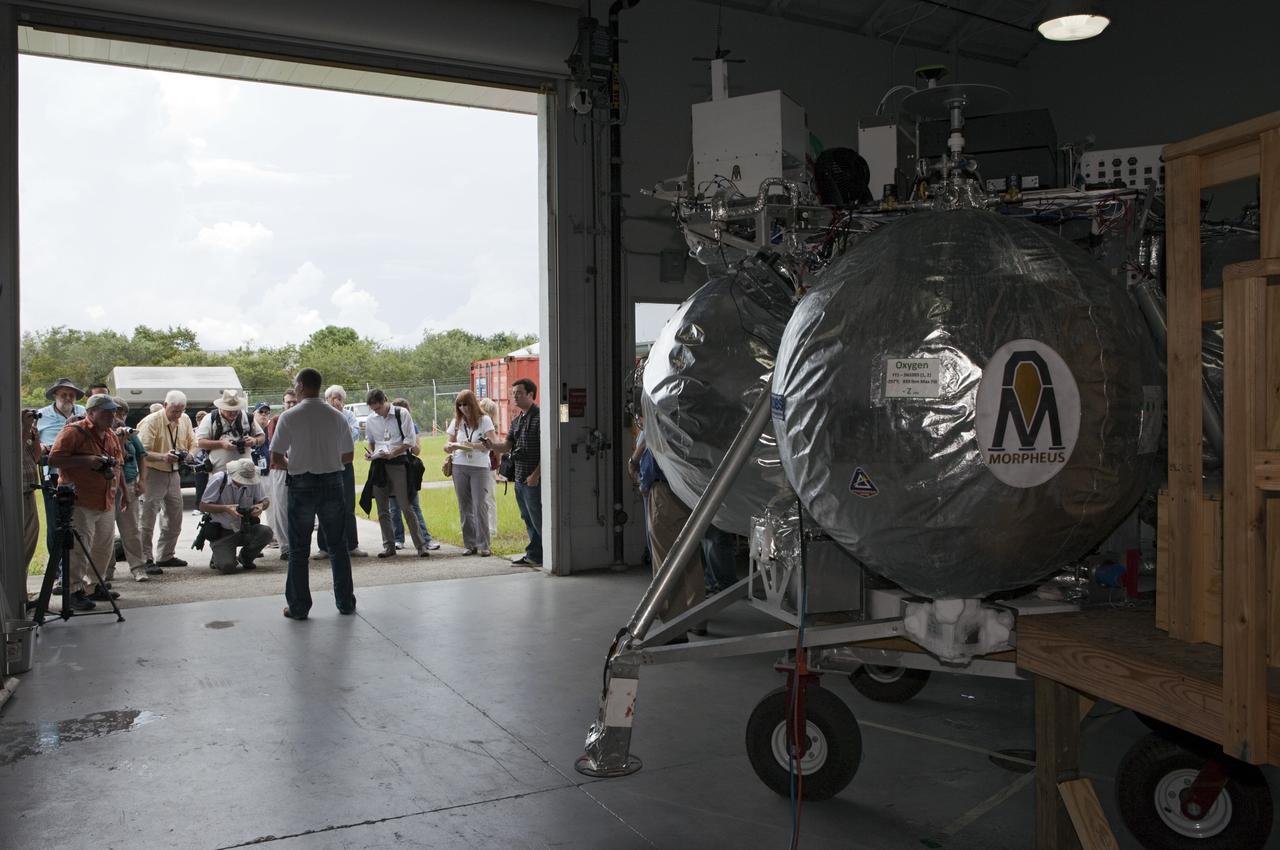 CAPE CANAVERAL, Fla. - At a hangar near the Shuttle Landing Facility, or SLF, at NASA’s Kennedy Space Center in Florida, the Johnson Space Center Project Morpheus Manager Jon Olansen speaks to members of the media. In the foreground is the Morpheus prototype lander, which arrived at Kennedy on July 27. Testing of the prototype lander had been ongoing at NASA’s Johnson Space Center in Houston in preparation for its first free-flight test at Kennedy Space Center. The SLF will provide the lander with the kind of field necessary for realistic testing, complete with rocks, craters and hazards to avoid. Morpheus utilizes an autonomous landing and hazard avoidance technology, or ALHAT, payload that will allow it to navigate to clear landing sites amidst rocks, craters and other hazards during its descent. Project Morpheus is one of 20 small projects comprising the Advanced Exploration Systems, or AES, program in NASA’s Human Exploration and Operations Mission Directorate. AES projects pioneer new approaches for rapidly developing prototype systems, demonstrating key capabilities and validating operational concepts for future human missions beyond Earth orbit. For more information on Project Morpheus, visit http://morpheuslander.jsc.nasa.gov/. Photo credit: NASA/Kim Shiflett