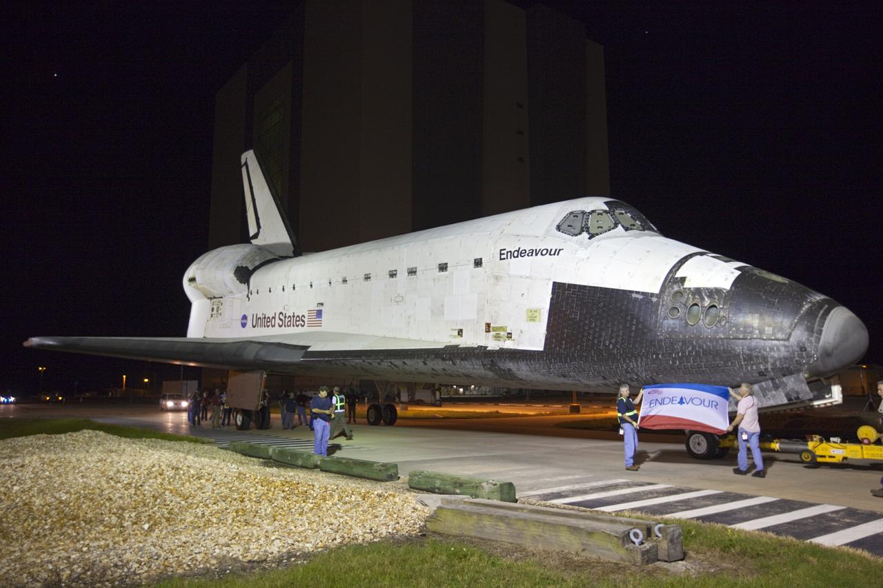 CAPE CANAVERAL, Fla. – While space shuttle Endeavour rolls toward the Shuttle Landing Facility, employees at NASA's Kennedy Space Center in Florida hold up a flag bearing the spacecraft's name. Endeavour moved to the nearby Shuttle Landing Facility, where it will be lifted in the gantry-like Mate-Demate Device and placed atop NASA's modified 747 Shuttle Carrier Aircraft for the final ferry flight of the Space Shuttle Program. Endeavour will be placed on permanent public display at the California Science Center in Los Angeles. Photo credit: NASA/Kim Shiflett