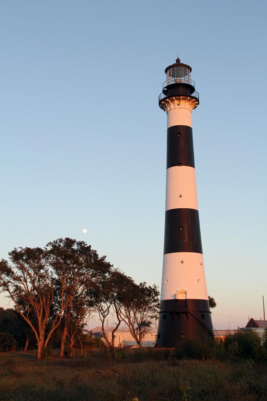 CAPE CANAVERAL AIR FORCE STATION, Fla. – The Cape Canaveral Air Force Station lighthouse takes on a warm glow as dawn breaks and a full moon still shines in the background. The Canaveral light is the only one owned by the U.S. Air Force.      In 2000, the Coast Guard transferred ownership of the lighthouse structure and its grounds to the Air Force, which is now responsible for maintaining it. The U.S. Coast Guard continues to operate the beacon as an active navigational aid. The first lighthouse at Cape Canaveral was built near the tip of the Cape in 1848. The structure was only about 60 feet high with a rather dim light powered by whale oil. In 1859, work began nearby on a new, taller iron structure. Construction was halted during the Civil War, and the lighthouse finally was finished in 1868. The structure, with a brick lining inside its iron exterior, was painted with its "daymark" black and white horizontal bands in 1873 to make it easier to identify during the day as a navigation point. Between 1892 and 1894, the lighthouse was dismantled and moved to its new home about a mile from the coast, where it stands today. Photo credit: NASA/Ben Smegelsky