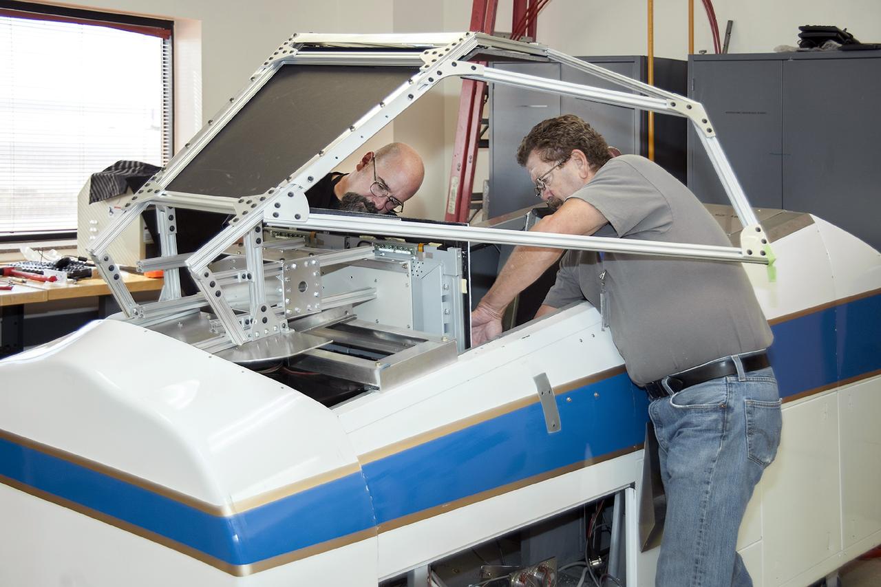 DRYDEN FLIGHT RESEARCH CENTER, Calif. - Simulation technicians Brent Bieber, left, and Dennis Pitts install a boilerplate Dream Chaser canopy structure over the cockpit of a flight simulator in the simulation laboratory at NASA's Dryden Flight Research Center in California. The modification will give Dream Chaser pilot-astronauts a more representative view of the actual flight profiles the spacecraft would fly during piloted approach and landing tests. Sierra Nevada Corporation's Space Systems division is conducting uncrewed captive- and free-flight approach and landing tests of its Dream Chaser at Dryden during the summer and fall. Photo credit: NASA/Ken Ulbrich