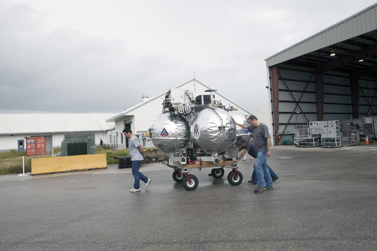 CAPE CANAVERAL, Fla. -- At NASA’s Kennedy Space Center in Florida, technicians move the Project Morpheus lander on a small transporter to a support building at the Shuttle Landing Facility, or SLF. Testing of the prototype lander has been ongoing at NASA’s Johnson Space Center in Houston in preparation for free flight testing at Kennedy. The SLF will provide the lander with the kind of field necessary for realistic testing, complete with rocks, craters and hazards to avoid. Morpheus utilizes an autonomous landing and hazard avoidance technology, or ALHAT, payload that will allow it to navigate to clear landing sites amidst rocks, craters and other hazards during its descent. Project Morpheus is one of 20 small projects comprising the Advanced Exploration Systems, or AES, program in NASA’s Human Exploration and Operations Mission Directorate. AES projects pioneer new approaches for rapidly developing prototype systems, demonstrating key capabilities and validating operational concepts for future human missions beyond Earth orbit. For more information on Project Morpheus, visit http://www.nasa.gov/centers/johnson/exploration/morpheus/index.html. Photo credit: NASA/Kim Shiflett