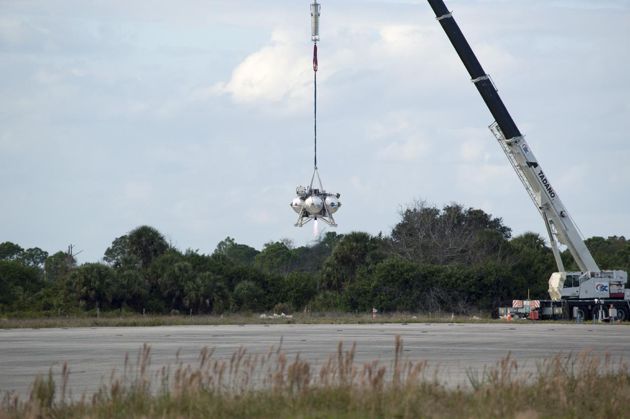 CAPE CANAVERAL, Fla. – At NASA’s Kennedy Space Center in Florida, the Project Morpheus prototype lander has been lifted by a tether and hovers above a transportable launch platform positioned at the north end of the Shuttle Landing Facility. The lander’s engine begins firing for a tethered test that includes lifting it 20 feet by crane, ascending another 10 feet, maneuvering backwards 10 feet, and then flying forward and descending to its original position, landing at the end of the tether. Testing of the prototype lander was performed at NASA’s Johnson Space Center in Houston in preparation for tethered and free flight testing at Kennedy. The landing facility will provide the lander with the kind of field necessary for realistic testing, complete with rocks, craters and hazards to avoid. Morpheus utilizes an autonomous landing and hazard avoidance technology, or ALHAT, payload that will allow it to navigate to clear landing sites amidst rocks, craters and other hazards during its descent. Project Morpheus is being managed under the Advanced Exploration Systems, or AES, Division in NASA’s Human Exploration and Operations Mission Directorate. The efforts in AES pioneer new approaches for rapidly developing prototype systems, demonstrating key capabilities and validating operational concepts for future human missions beyond Earth orbit. For more information on Project Morpheus, visit http://morpheuslander.jsc.nasa.gov. Photo credit: NASA/Daniel Casper