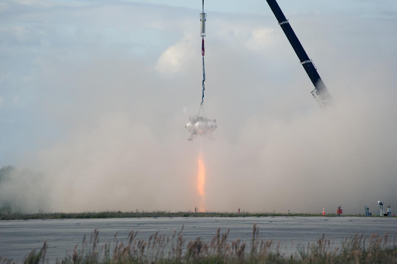 CAPE CANAVERAL, Fla. – At NASA’s Kennedy Space Center in Florida, smoke fills the air as the Project Morpheus prototype lander’s engine fires during a tether test at the north end of the Shuttle Landing Facility. During the test, the lander was lifted 20 feet by crane, and then ascended another 10 feet. The lander will maneuver backwards 10 feet, and then fly forward and descend to its original position, landing at the end of the tether onto a transportable launch platform. Testing of the prototype lander was performed at NASA’s Johnson Space Center in Houston in preparation for tethered and free flight testing at Kennedy.    The landing facility will provide the lander with the kind of field necessary for realistic testing, complete with rocks, craters and hazards to avoid. Morpheus utilizes an autonomous landing and hazard avoidance technology, or ALHAT, payload that will allow it to navigate to clear landing sites amidst rocks, craters and other hazards during its descent. Project Morpheus is being managed under the Advanced Exploration Systems, or AES, Division in NASA’s Human Exploration and Operations Mission Directorate. The efforts in AES pioneer new approaches for rapidly developing prototype systems, demonstrating key capabilities and validating operational concepts for future human missions beyond Earth orbit. For more information on Project Morpheus, visit http://morpheuslander.jsc.nasa.gov.  Photo credit: NASA/Daniel Casper