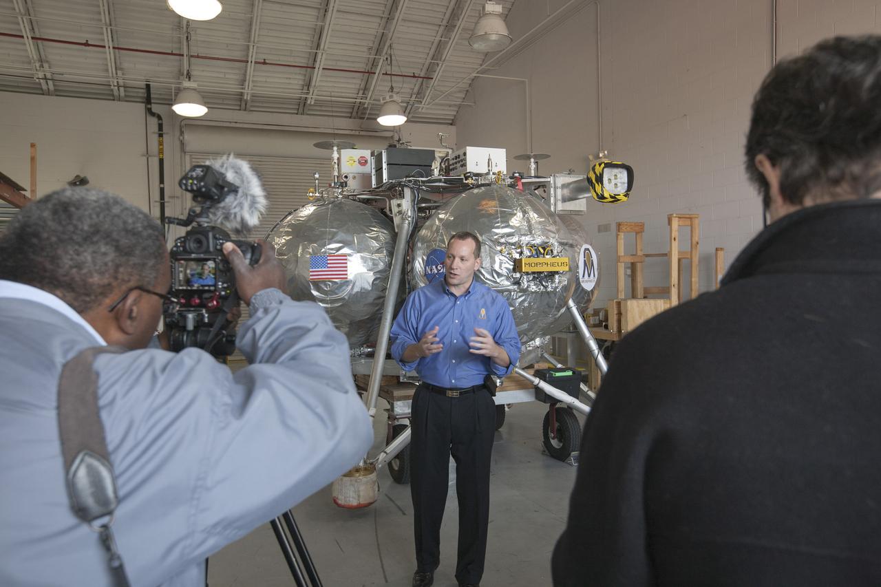 CAPE CANAVERAL, Fla. – Members of the news media view the Project Morpheus prototype lander inside a hangar near the Shuttle Landing Facility at NASA’s Kennedy Space Center in Florida. Speaking to the media is Jon Olansen, the Morpheus project manager at Johnson Space Center in Houston. Morpheus successfully completed its third free flight test Jan. 16. The 57-second test began at 1:15 p.m. EST with the Morpheus lander launching from the ground over a flame trench and ascending about 187 feet, nearly doubling the target ascent velocity from the last test in December 2013. The lander flew forward, covering about 154 feet in 20 seconds before descending and landing within 11 inches of its target on a dedicated pad inside the autonomous landing and hazard avoidance technology, or ALHAT, hazard field. Project Morpheus tests NASA’s ALHAT and an engine that runs on liquid oxygen and methane, or green propellants, into a fully-operational lander that could deliver cargo to other planetary surfaces. The landing facility provides the lander with the kind of field necessary for realistic testing, complete with rocks, craters and hazards to avoid. Morpheus’ ALHAT payload allows it to navigate to clear landing sites amidst rocks, craters and other hazards during its descent. Project Morpheus is being managed under the Advanced Exploration Systems, or AES, Division in NASA’s Human Exploration and Operations Mission Directorate. The efforts in AES pioneer new approaches for rapidly developing prototype systems, demonstrating key capabilities and validating operational concepts for future human missions beyond Earth orbit. For more information on Project Morpheus, visit http://www.nasa.gov/centers/johnson/exploration/morpheus. Photo credit: NASA/Kim Shiflett