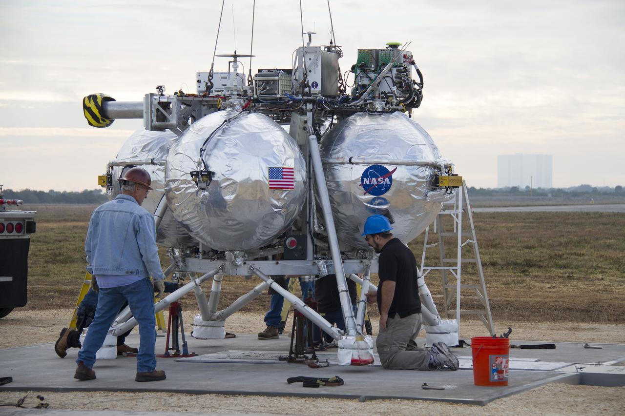 CAPE CANAVERAL, Fla. – Technicians monitor the progress as a crane lowers the Project Morpheus prototype for positioning on a launch pad at the north end of the Shuttle Landing Facility at NASA’s Kennedy Space Center in Florida. The prototype lander is being prepared for its fourth free flight test at Kennedy. Morpheus will launch from the ground over a flame trench and then descend and land on a dedicated pad inside the autonomous landing and hazard avoidance technology, or ALHAT, hazard field. Project Morpheus tests NASA’s ALHAT and an engine that runs on liquid oxygen and methane, or green propellants, into a fully-operational lander that could deliver cargo to other planetary surfaces. The landing facility provides the lander with the kind of field necessary for realistic testing, complete with rocks, craters and hazards to avoid. Morpheus’ ALHAT payload allows it to navigate to clear landing sites amidst rocks, craters and other hazards during its descent. Project Morpheus is being managed under the Advanced Exploration Systems, or AES, Division in NASA’s Human Exploration and Operations Mission Directorate. The efforts in AES pioneer new approaches for rapidly developing prototype systems, demonstrating key capabilities and validating operational concepts for future human missions beyond Earth orbit. For more information on Project Morpheus, visit http://www.nasa.gov/centers/johnson/exploration/morpheus. Photo credit: NASA/Cory Huston