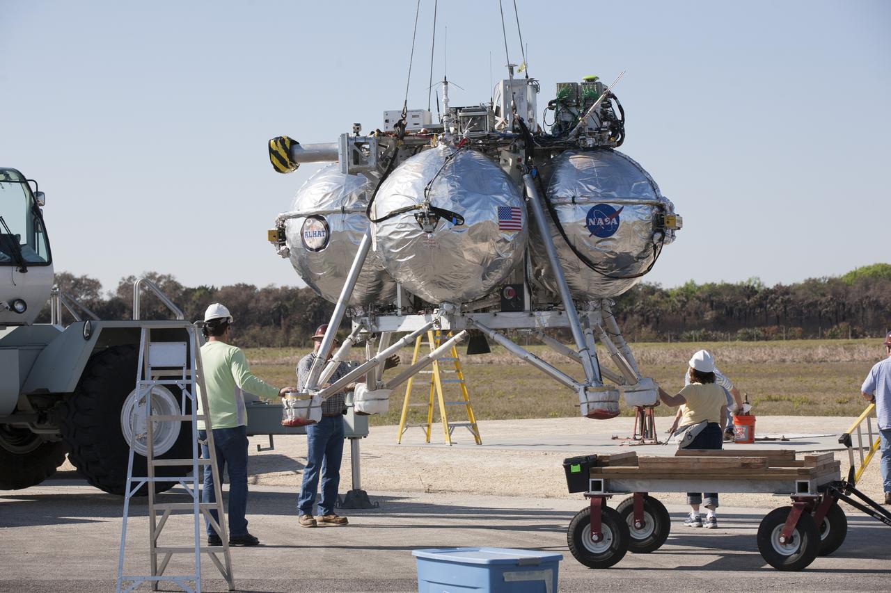 CAPE CANAVERAL, Fla. - Engineers and technicians assist as a crane lowers the Project Morpheus prototype lander in preparation for its seventh free flight test at the north end of the Shuttle Landing Facility at NASA’s Kennedy Space Center in Florida. The 83-second test began at 3:41 p.m. EDT with the Morpheus lander launching from the ground over a flame trench and ascending to 580 feet, its highest to date. Morpheus then flew its fastest downrange trek at 30 mph, travelling farther than before, 837 feet. The lander performed a 42-foot divert to emulate a hazard avoidance maneuver before descending and touching down on Landing Site 2, at the northern landing pad inside the automated landing and hazard avoidance technology ALHAT hazard field. Morpheus landed within one foot of its intended target. Project Morpheus tests NASA’s ALHAT and an engine that runs on liquid oxygen and methane, or green propellants, into a fully-operational lander that could deliver cargo to other planetary surfaces. The landing facility provides the lander with the kind of field necessary for realistic testing, complete with rocks, craters and hazards to avoid. Morpheus’ ALHAT payload allows it to navigate to clear landing sites amidst rocks, craters and other hazards during its descent. Project Morpheus is being managed under the Advanced Exploration Systems, or AES, Division in NASA’s Human Exploration and Operations Mission Directorate. The efforts in AES pioneer new approaches for rapidly developing prototype systems, demonstrating key capabilities and validating operational concepts for future human missions beyond Earth orbit. For more information on Project Morpheus, visit http://morpheuslander.jsc.nasa.gov/. Photo credit: NASA/Kim Shiflett