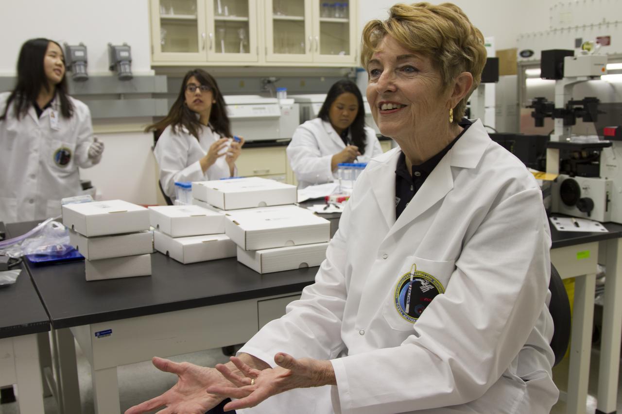 CAPE CANAVERAL, Fla. - Researcher and principal investigator Dr. Millie Hughes-Fulford, of the Hughes-Fulford Laboratory, San Francisco, Calif., discusses her laboratory's T-cell experiment and the impact the research may have on aging adults and their immune systems with an interviewer in the Space Station Processing Facility. From left, T-cell science team members Miya Yoshida, Emily Martinez and Tara Candelario are at work preparing for launch in the background. The immunology experiment will launch on SpaceX-3 and focus on the effects of microgravity on early T-cell signaling pathways. Current work aims to identify and compare the gene expression of microRNAs miRNAs during T-cell activation under normal gravity and in microgravity, and compare those patterns to changes seen in aging populations. The experiment will be the first flown on SpaceX funded by the National Institutes of Health. Dr. Hughes-Fulford flew aboard space shuttle mission STS-40 in June 1991, the first Spacelab mission dedicated to biomedical studies. For more information on the T-cell experiment, visit http://hughesfulfordlab.com and http://www.nasa.gov/ames/research/space-biosciences/t-cell-activation-in-aging-spacex-3/. Photo credit: NASA/Cory Huston