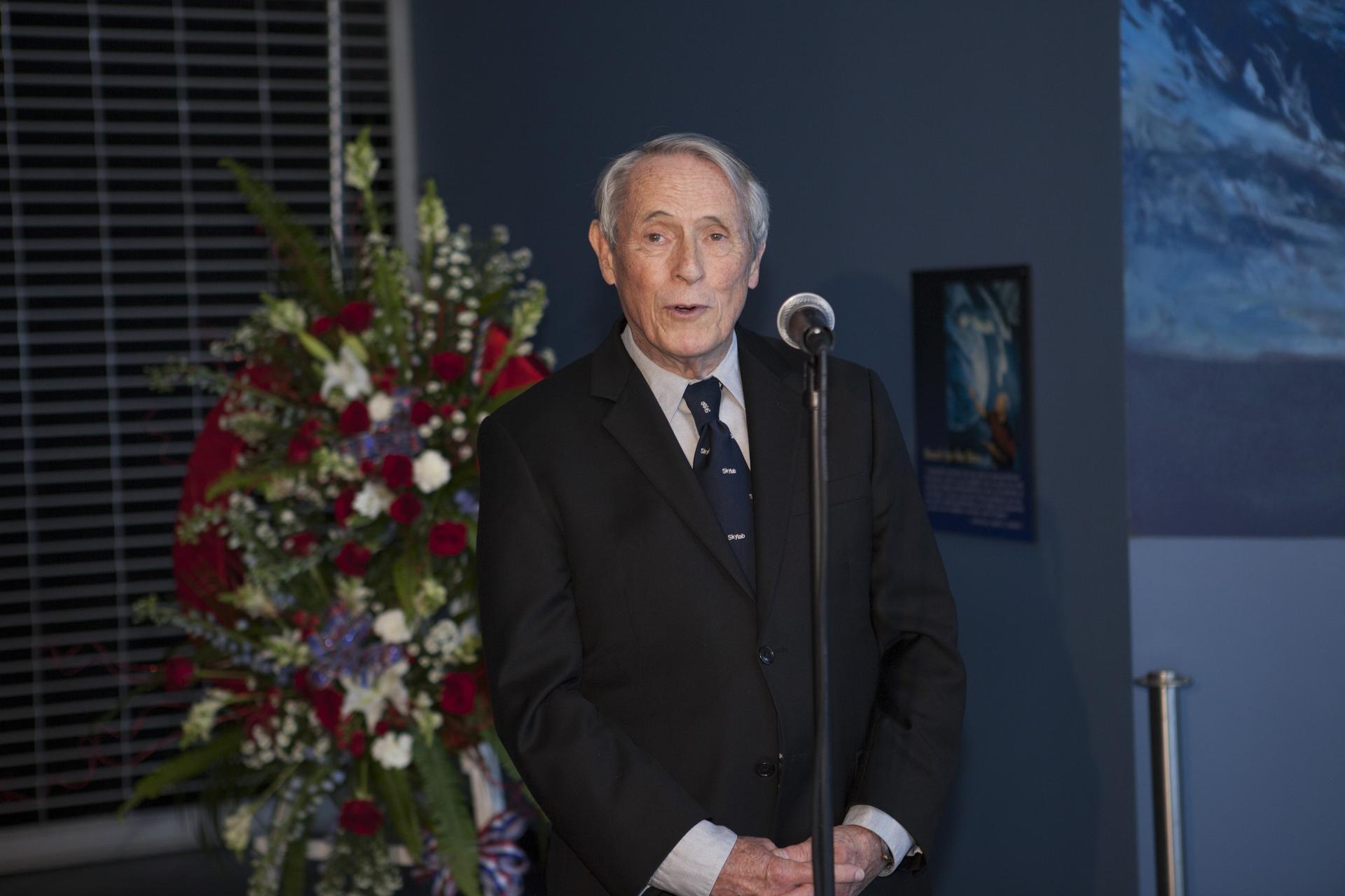 Former astronaut Ed Gibson speaks into a microphone. A large wreath is behind him.