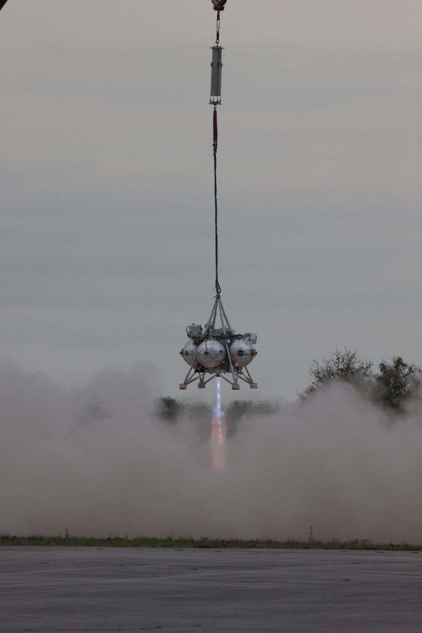 CAPE CANAVERAL, Fla. – NASA's Project Morpheus prototype lander is lifted by crane and its engine is ignited during a tethered test near a new launch site at the north end of the Shuttle Landing Facility at NASA's Kennedy Space Center in Florida. The launch pad was moved to a different location at the landing facility to support the next phase of flight testing. The test was performed to verify the lander's recently installed autonomous landing and hazard avoidance technology, or ALHAT, sensors and integration systems. During the test, Morpheus was lifted by crane to 20 feet for an engine firing of about 74 seconds and then lowered to the ground. With the successful completion of the test, the Morpheus project team will begin preparing for the first free flight test with ALHAT. Morpheus tests NASA’s ALHAT, and an engine that runs on liquid oxygen and methane, or green propellants, into a fully-operational lander that could deliver cargo to other planetary surfaces. The landing facility provides the lander with the kind of field necessary for realistic testing, complete with rocks, craters and hazards to avoid. Morpheus’ ALHAT payload allows it to navigate to clear landing sites amidst rocks, craters and other hazards during its descent. Project Morpheus is being managed under the Advanced Exploration Systems, or AES, Division in NASA’s Human Exploration and Operations Mission Directorate. The efforts in AES pioneer new approaches for rapidly developing prototype systems, demonstrating key capabilities and validating operational concepts for future human missions beyond Earth orbit. For more information on Project Morpheus, visit http://morpheuslander.jsc.nasa.gov/. Photo credit: NASA/Glenn Benson