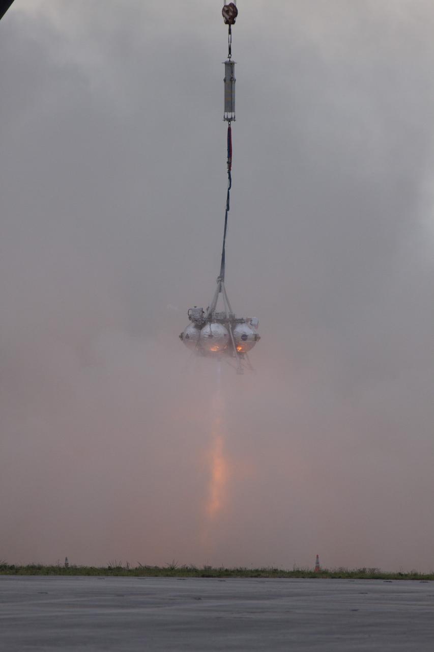 CAPE CANAVERAL, Fla. – NASA's Project Morpheus prototype lander is lifted by crane and its engine is ignited during a tethered test near a new launch site at the north end of the Shuttle Landing Facility at NASA's Kennedy Space Center in Florida. The launch pad was moved to a different location at the landing facility to support the next phase of flight testing. The test was performed to verify the lander's recently installed autonomous landing and hazard avoidance technology, or ALHAT, sensors and integration systems. During the test, Morpheus was lifted by crane to 20 feet for an engine firing of about 74 seconds and then lowered to the ground. With the successful completion of the test, the Morpheus project team will begin preparing for the first free flight test with ALHAT. Morpheus tests NASA’s ALHAT, and an engine that runs on liquid oxygen and methane, or green propellants, into a fully-operational lander that could deliver cargo to other planetary surfaces. The landing facility provides the lander with the kind of field necessary for realistic testing, complete with rocks, craters and hazards to avoid. Morpheus’ ALHAT payload allows it to navigate to clear landing sites amidst rocks, craters and other hazards during its descent. Project Morpheus is being managed under the Advanced Exploration Systems, or AES, Division in NASA’s Human Exploration and Operations Mission Directorate. The efforts in AES pioneer new approaches for rapidly developing prototype systems, demonstrating key capabilities and validating operational concepts for future human missions beyond Earth orbit. For more information on Project Morpheus, visit http://morpheuslander.jsc.nasa.gov/. Photo credit: NASA/Glenn Benson