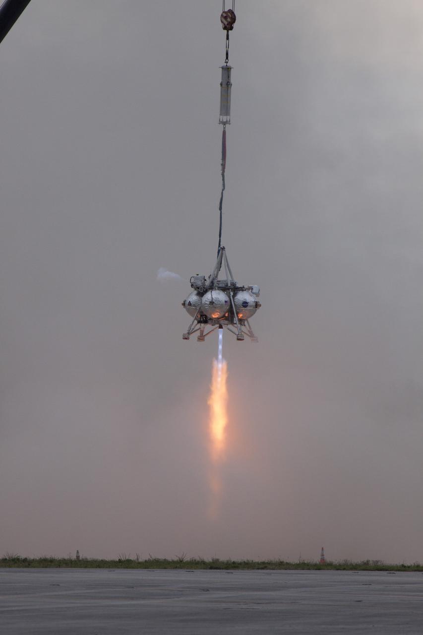 CAPE CANAVERAL, Fla. – NASA's Project Morpheus prototype lander is lifted by crane and its engine is ignited during a tethered test near a new launch site at the north end of the Shuttle Landing Facility at NASA's Kennedy Space Center in Florida. The launch pad was moved to a different location at the landing facility to support the next phase of flight testing. The test was performed to verify the lander's recently installed autonomous landing and hazard avoidance technology, or ALHAT, sensors and integration systems. During the test, Morpheus was lifted by crane to 20 feet for an engine firing of about 74 seconds and then lowered to the ground. With the successful completion of the test, the Morpheus project team will begin preparing for the first free flight test with ALHAT. Morpheus tests NASA’s ALHAT, and an engine that runs on liquid oxygen and methane, or green propellants, into a fully-operational lander that could deliver cargo to other planetary surfaces. The landing facility provides the lander with the kind of field necessary for realistic testing, complete with rocks, craters and hazards to avoid. Morpheus’ ALHAT payload allows it to navigate to clear landing sites amidst rocks, craters and other hazards during its descent. Project Morpheus is being managed under the Advanced Exploration Systems, or AES, Division in NASA’s Human Exploration and Operations Mission Directorate. The efforts in AES pioneer new approaches for rapidly developing prototype systems, demonstrating key capabilities and validating operational concepts for future human missions beyond Earth orbit. For more information on Project Morpheus, visit http://morpheuslander.jsc.nasa.gov/. Photo credit: NASA/Glenn Benson