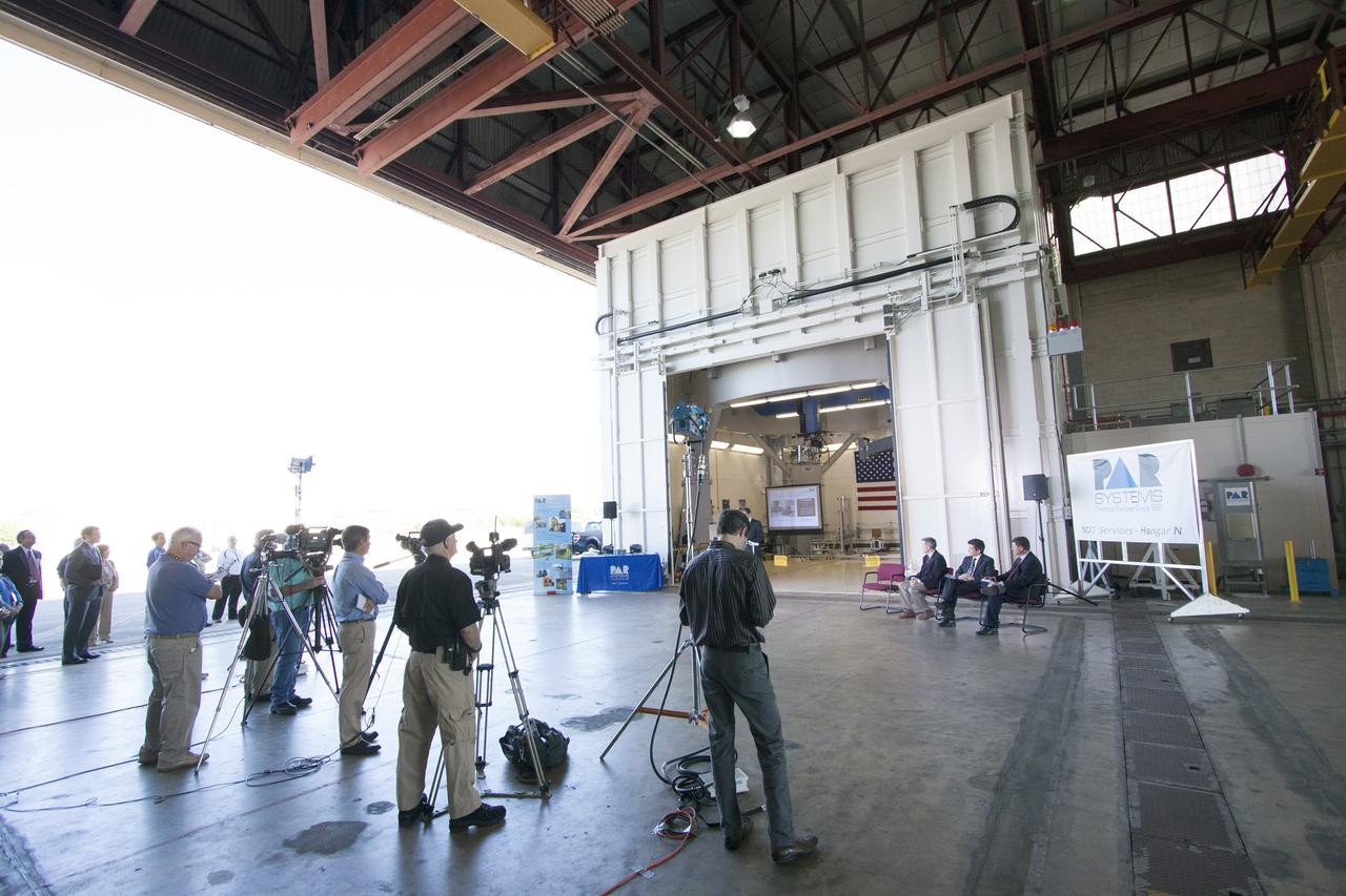 CAPE CANAVERAL, Fla. – Brian Behm partially hidden, president, aerospace robotics, PaR Systems Inc., speaks during an Open House event at Hangar N at Cape Canaveral Air Force Station in Florida, to celebrate the one-year anniversary of a partnership with NASA Kennedy Space Center. Under a 15-year lease agreement, PaR Systems is utilizing Hangar N and its unique nondestructive testing equipment. Behind Behm is the robotic inspection cell that contains an automated X-ray system once used to scan the aft skirts of the solid rocket boosters for the space shuttle.    The partnership agreement was established by Kennedy's Center Planning and Development Directorate. The agreement is just one example of the types of partnerships that Kennedy is seeking to create a multi-user spaceport.  Photo credit: NASA/Cory Huston