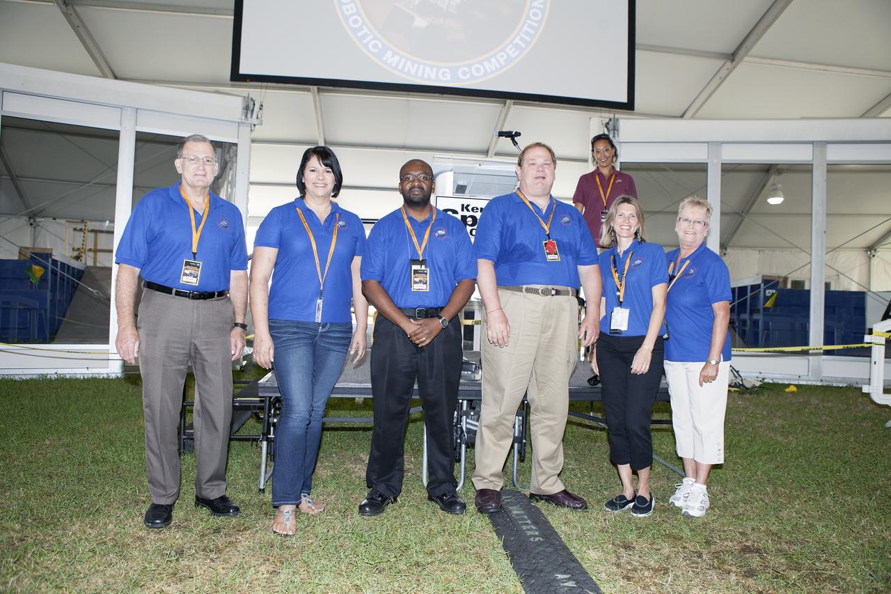 CAPE CANAVERAL, Fla. – The presentation and team spirit judges for NASA's 2014 Robotics Mining Competition are introduced during the opening ceremony at the Kennedy Space Center Visitor Complex in Florida. Second from left, is Teresa Martinez, lead presentation judge from Kennedy's Education Office.  At far right, is Beth Smith, lead team spirit judge from Kennedy's Education Office. Behind them on the podium is Kimberley Land, event emcee from NASA's Ames Research Center in Moffett Field, California. More than 35 teams from around the U.S. have designed and built remote-controlled robots for the mining competition.    The competition is a NASA Human Exploration and Operations Mission Directorate project designed to engage and retain students in science, technology, engineering and mathematics, or STEM, fields by expanding opportunities for student research and design. Teams use their remote-controlled robotics to maneuver and dig in a supersized sandbox filled with a crushed material that has characteristics similar to Martian soil. The objective of the challenge is to see which team’s robot can collect and move the most regolith within a specified amount of time. For more information, visit www.nasa.gov/nasarmc. Photo credit: NASA/Frankie Martin