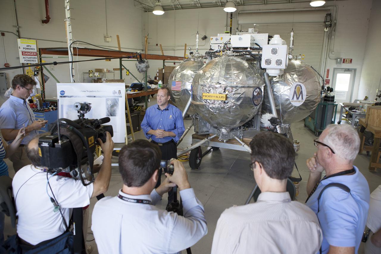 CAPE CANAVERAL, Fla. – Jon Olansen, Morpheus project manager, speaks to members of the media inside a facility near the Shuttle Landing Facility at NASA's Kennedy Space Center in Florida. Behind Olansen is the Project Morpheus prototype lander. Project Morpheus tests NASA’s autonomous landing and hazard avoidance technology, or ALHAT, sensors and an engine that runs on liquid oxygen and methane, which are green propellants. These new capabilities could be used in future efforts to deliver cargo to planetary surfaces.    The landing facility provides the lander with the kind of field necessary for realistic testing, complete with rocks, craters and hazards to avoid. Morpheus’ ALHAT payload allows it to navigate to clear landing sites amidst rocks, craters and other hazards during its descent. Project Morpheus is being managed under the Advanced Exploration Systems, or AES, Division in NASA’s Human Exploration and Operations Mission Directorate. The efforts in AES pioneer new approaches for rapidly developing prototype systems, demonstrating key capabilities and validating operational concepts for future human missions beyond Earth orbit. For more information on Project Morpheus, visit http://morpheuslander.jsc.nasa.gov/.  Photo credit: NASA/Frankie Martin