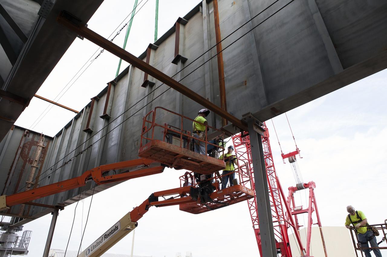 CAPE CANAVERAL, Fla. – Modifications continue on the Mobile Launcher, or ML, at the Mobile Launcher Park Site at NASA's Kennedy Space Center in Florida. Construction workers on lifts watch as a crane is used to bring the final large steel beam closer for installation on the base of the ML. The ML is being modified and strengthened to accommodate the weight, size and thrust at launch of NASA's Space Launch System, or SLS, and Orion spacecraft.    In 2013, the agency awarded a contract to J.P. Donovan Construction Inc. of Rockledge, Fla., to modify the ML, which is one of the key elements of ground support equipment that is being upgraded by the Ground Systems Development and Operations Program at Kennedy. The existing 24-foot exhaust hole is being enlarged and strengthened for the larger, heavier SLS rocket. The ML will carry the SLS rocket and Orion spacecraft to Launch Pad 39B for its first uncrewed mission, Exploration Mission-1, in 2018. Photo credit: NASA/Daniel Casper