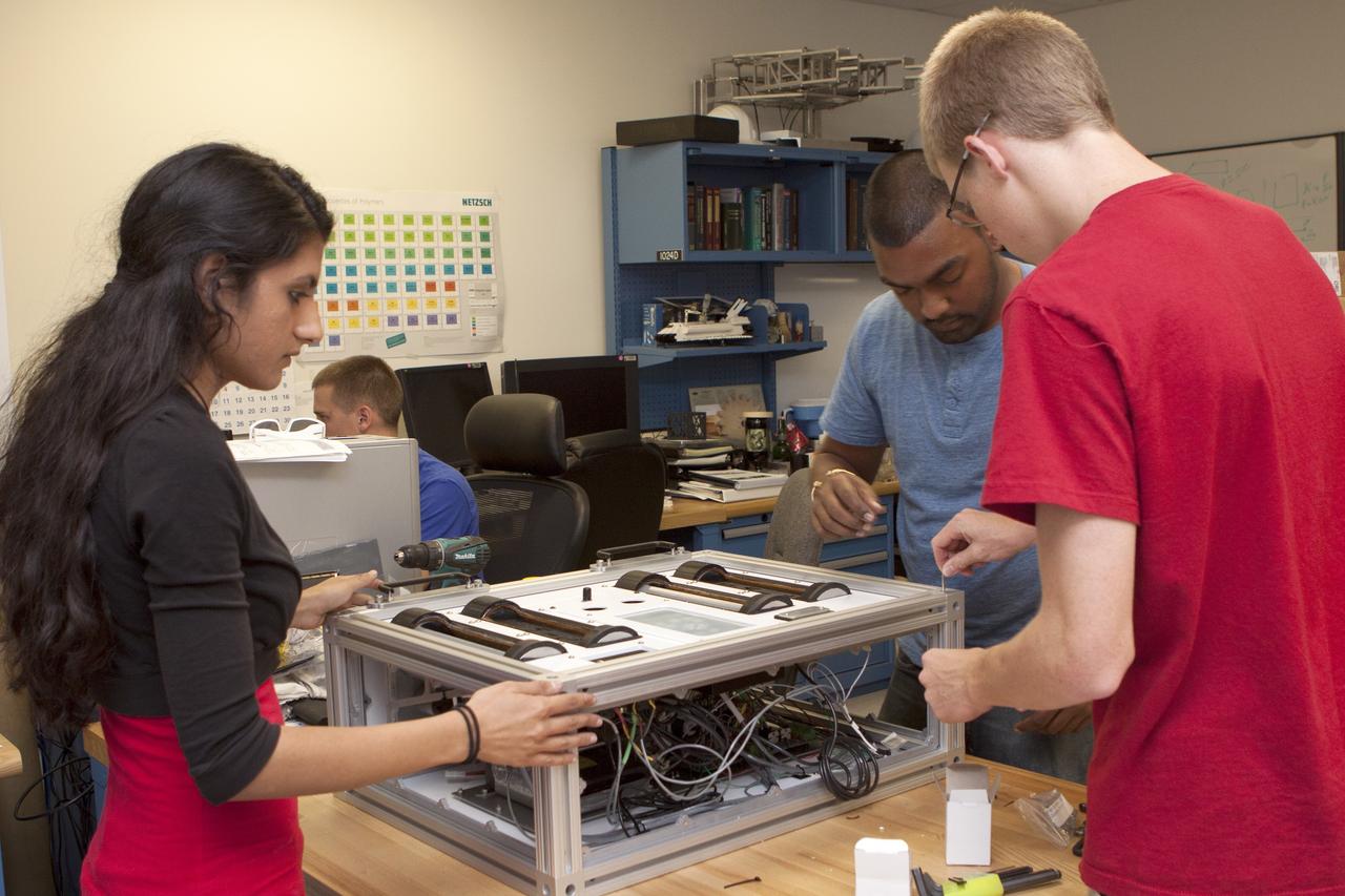 CAPE CANAVERAL, Fla. – Inside the Prototype Laboratory at NASA's Kennedy Space Center in Florida, Prital Thakrar, left, design lead and student engineer trainee from the University of Florida in Gainesville, Anthony Bharrat, NASA avionics lead, and Evan Williams, an Education intern from the University of Central Florida, prepare the experiment container for NASA's Exposing Microorganisms in the Stratosphere, or E-MIST, experiment. The container was designed and built at Kennedy. The 80-pound structure features four doors that rotate to expose up to 10 microbial samples each for a predetermined period of time in the Earth's stratosphere.    The E-MIST experiment will launch on the exterior of a giant scientific balloon gondola at about 8 a.m. MST on Aug. 24 from Ft. Sumner, New Mexico. It will soar 125,000 feet above the Earth during a 5-hour journey over the desert to understand how spore-forming bacteria, commonly found in spacecraft assembly facilities can survive. Photo credit: NASA/Kim Shiflett